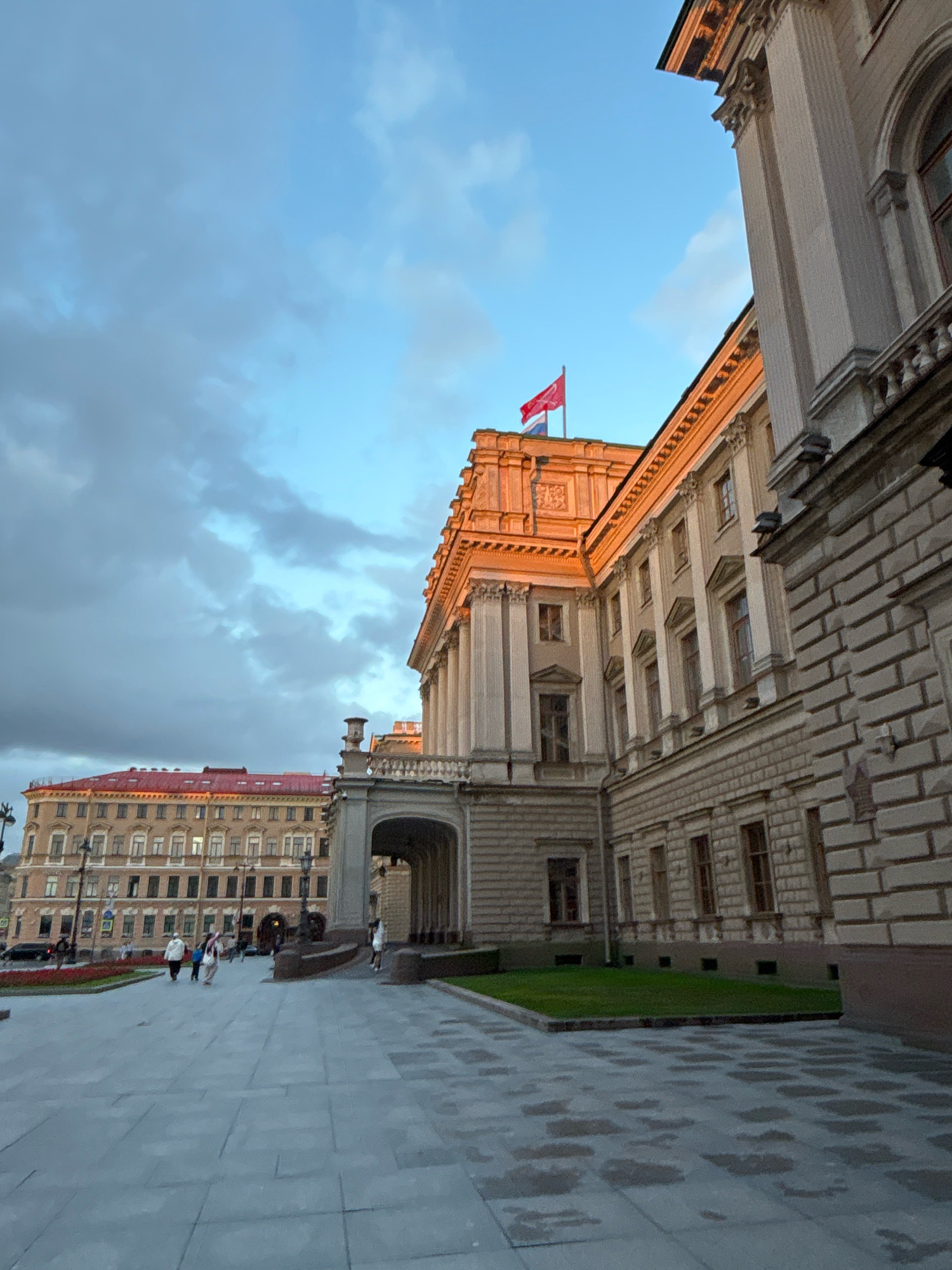 The image shows a view of the Palace Square in Saint Petersburg, Russia, at dusk. The sun is setting, casting a warm glow on the buildings. In the foreground, a paved square with wet patches suggests recent rain. People are walking on the square. In the background, the Winter Palace, a grand baroque building, dominates the scene. The sky is partly cloudy with a gradient of blue and grey hues. A red flag is visible atop one of the buildings.