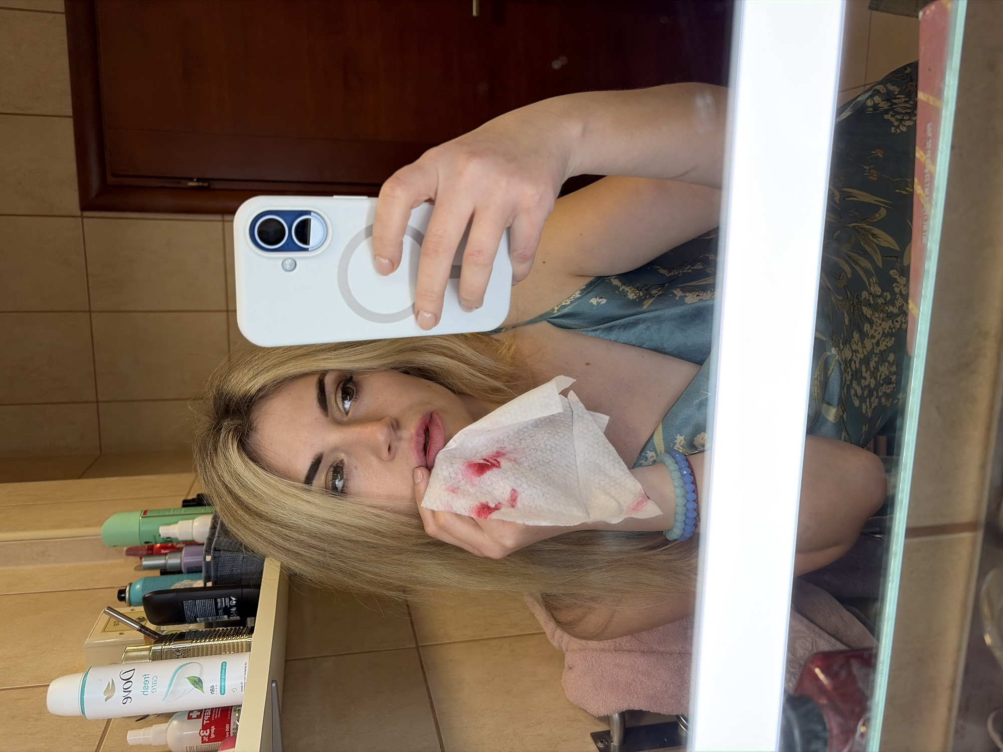 A blonde female is depicted in a mirror selfie within an indoor bathroom setting. She wears a teal slip dress with a light floral pattern and two beaded bracelets on her left wrist. Her right hand holds a white, textured tissue, heavily stained with a significant quantity of red material, pressed against her lips, which also appear reddened. She holds a white smartphone with visible dual cameras and a rear magnetic ring feature in her left hand, positioned to capture her reflection. Her expression is serious, and her gaze is directed towards the camera. The background features light brown tiled walls, a wooden shelf containing various toiletries including a Dove Fresh Care deodorant spray, and a dark brown wooden door on the right. A pink towel is reflected below the visible mirror edge. The scene is illuminated by a bright light strip positioned along the top edge of the mirror. While the environment is consistent with a private or hotel bathroom, the specific location within Budva, Montenegro, cannot be determined from the visual information provided in the image.