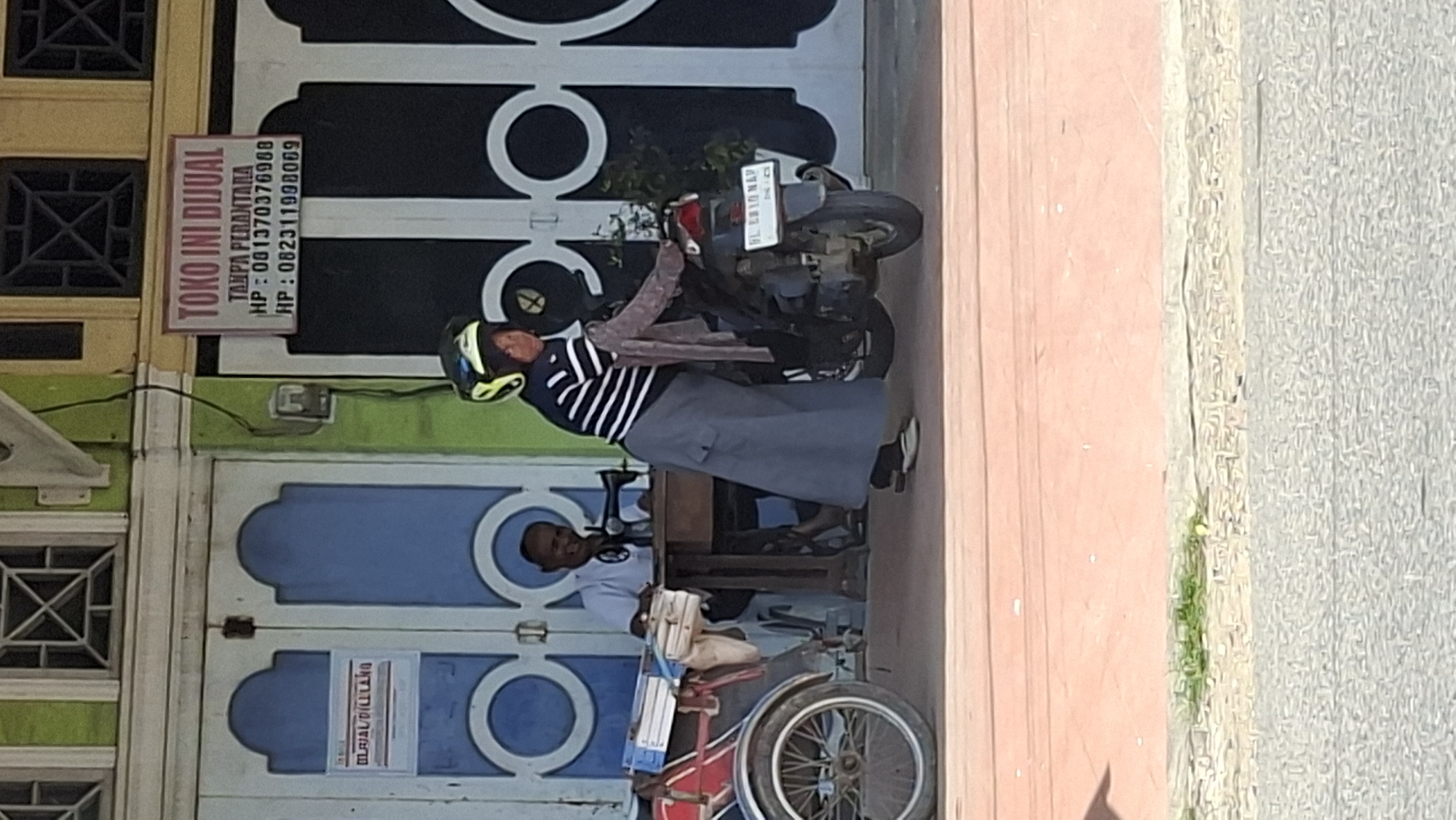 In front of a building with a green and yellow facade in Lhokseumawe, Indonesia, a man sits behind a table with a sewing machine. A woman, wearing a striped shirt, grey skirt, and a helmet, is standing next to a motorcycle. A sign on the building advertises "TOKO INI DIJUAL" (This Shop is For Sale). A tricycle with books is parked to the left of the table. The ground in front of the building is paved with red bricks, and a road is visible beyond.