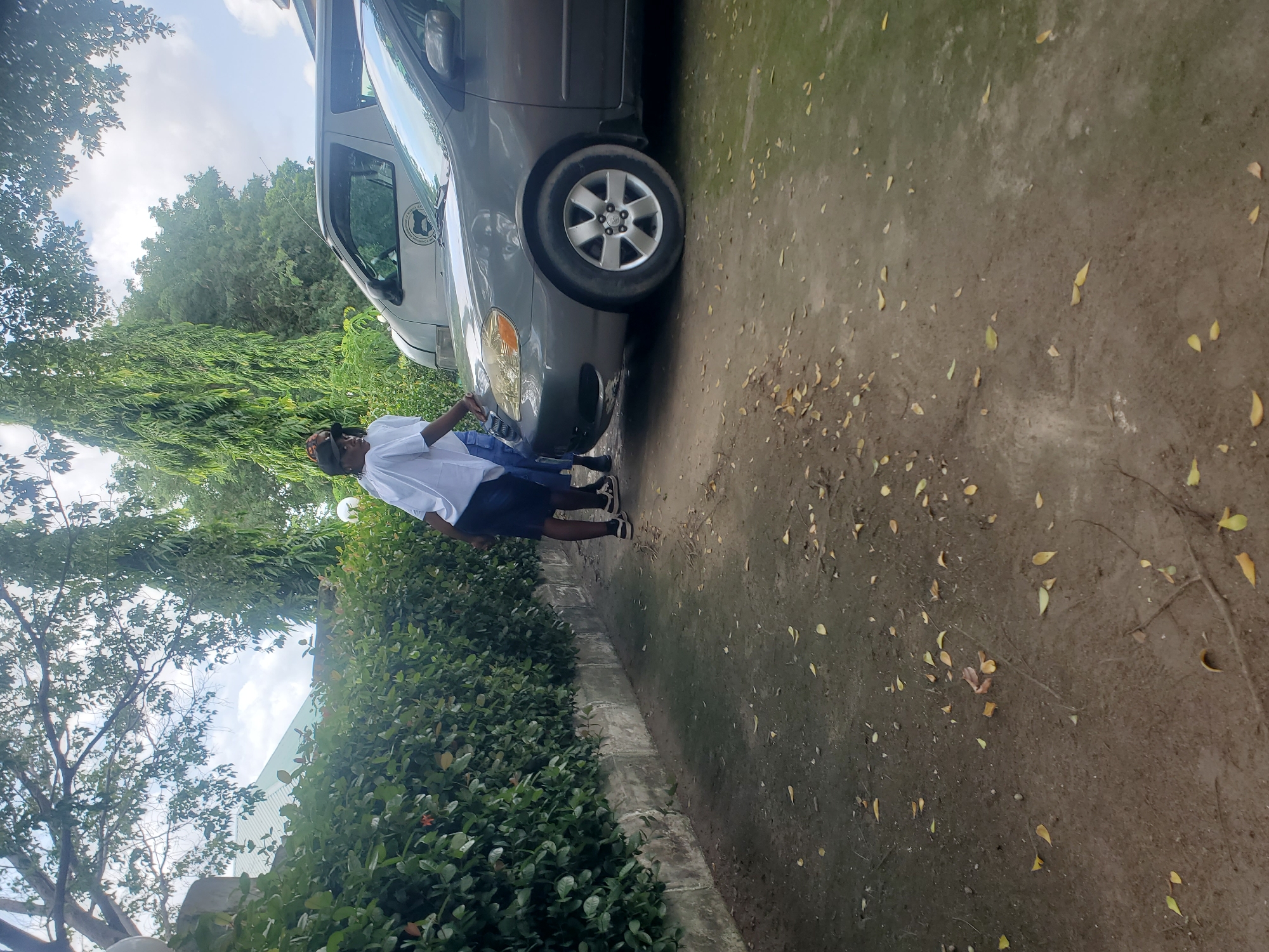 A person, likely a young male, is standing next to a gray car. The person is wearing a white t-shirt and dark shorts. The scene is outdoors with a lot of greenery from trees and bushes. The ground appears to be dirt or gravel, with fallen leaves scattered around. The sky is visible in the background, suggesting it's daytime. The car is parked on what looks like a driveway or a side road. The image appears to be taken in Jalingo, Nigeria, as per the provided context. The person's stance suggests they might be waiting or observing something. There are no visible emotions conveyed by the person's posture or expression. The overall setting is casual and appears to be a warm day.