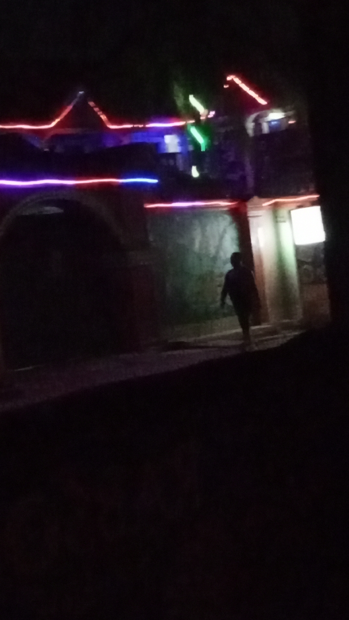 This nighttime image features a blurry, outdoor scene dominated by artificial lights and deep shadows. The primary subject is a person, visible as a dark silhouette, walking from left to right across the midground. Their posture suggests movement, but details of their clothing or features are indistinct.
In the background, a building is illuminated by numerous colorful string lights, predominantly red, purple, blue, and green. These lights trace the outline of the building's roof and possibly lower architectural elements, creating a festive or decorative atmosphere. An arched doorway or entrance, heavily shadowed, occupies the left side of the frame, suggesting a distinct architectural style. To the right of the walking figure, a wall or structure is faintly lit with a greenish hue, and further right, a bright, rectangular light source, possibly a window or a sign, emits a white glow.
The setting is clearly at night, with most of the scene enveloped in darkness, highlighting the artificial illumination. The foreground is entirely obscured by shadow. No specific location cues are discernible beyond the architectural hints and the prevalence of string lights. There is no visible text in the image. The blurriness throughout the image suggests it was captured in low light conditions, possibly handheld, contributing to a sense of dynamism or spontaneity.