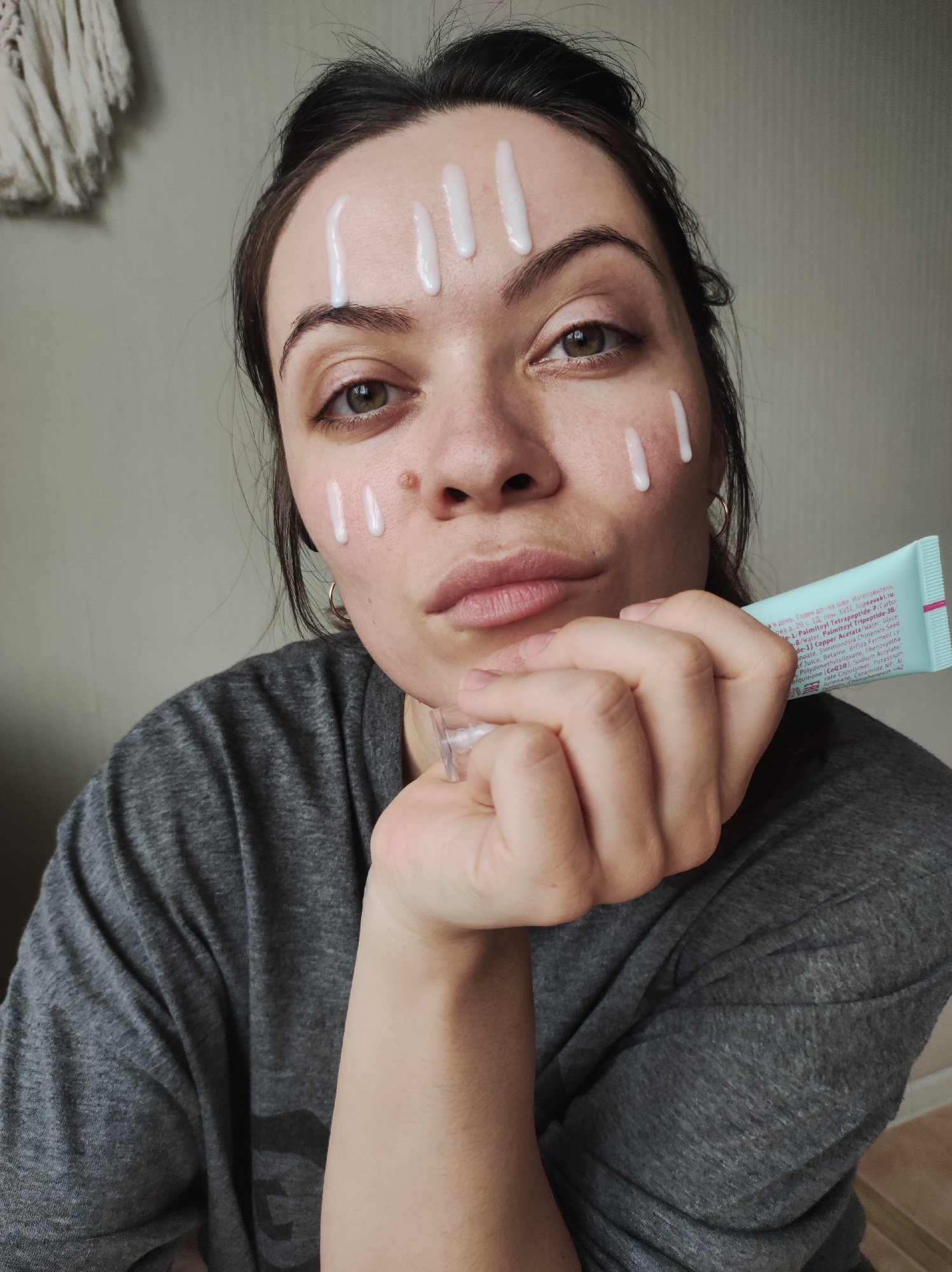 A woman is shown in a close-up shot, applying or having applied a cream or lotion to her face. Several distinct, vertical lines of white cream are visible on her forehead and cheeks. She is holding a tube of what appears to be the same cream in her hand. The tube has Cyrillic text printed on it. She's wearing a gray t-shirt, and her hair is pulled back loosely. Her expression is somewhat pensive or pouty. A decorative wall hanging is partially visible in the upper left corner of the frame. The background is a plain wall.