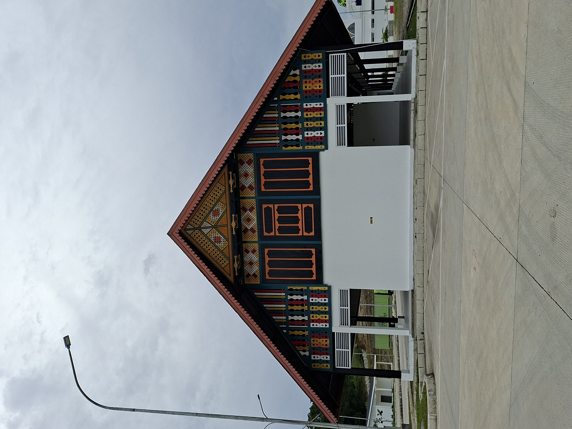 A striking building featuring traditional Acehnese architectural elements stands prominently in Keureuweungkrueng, Indonesia. The building's facade showcases a steeply pitched roof adorned with vibrant, geometric designs in hues of yellow, orange, red, black, and white. Below the roof, the building transitions to a modern white structure with vertical white slats and black structural elements.
The building is surrounded by a paved concrete area, likely a parking lot or plaza. In the foreground, a street light stands tall against a cloudy sky. The overall scene captures a blend of cultural heritage and contemporary design within a developing urban environment.