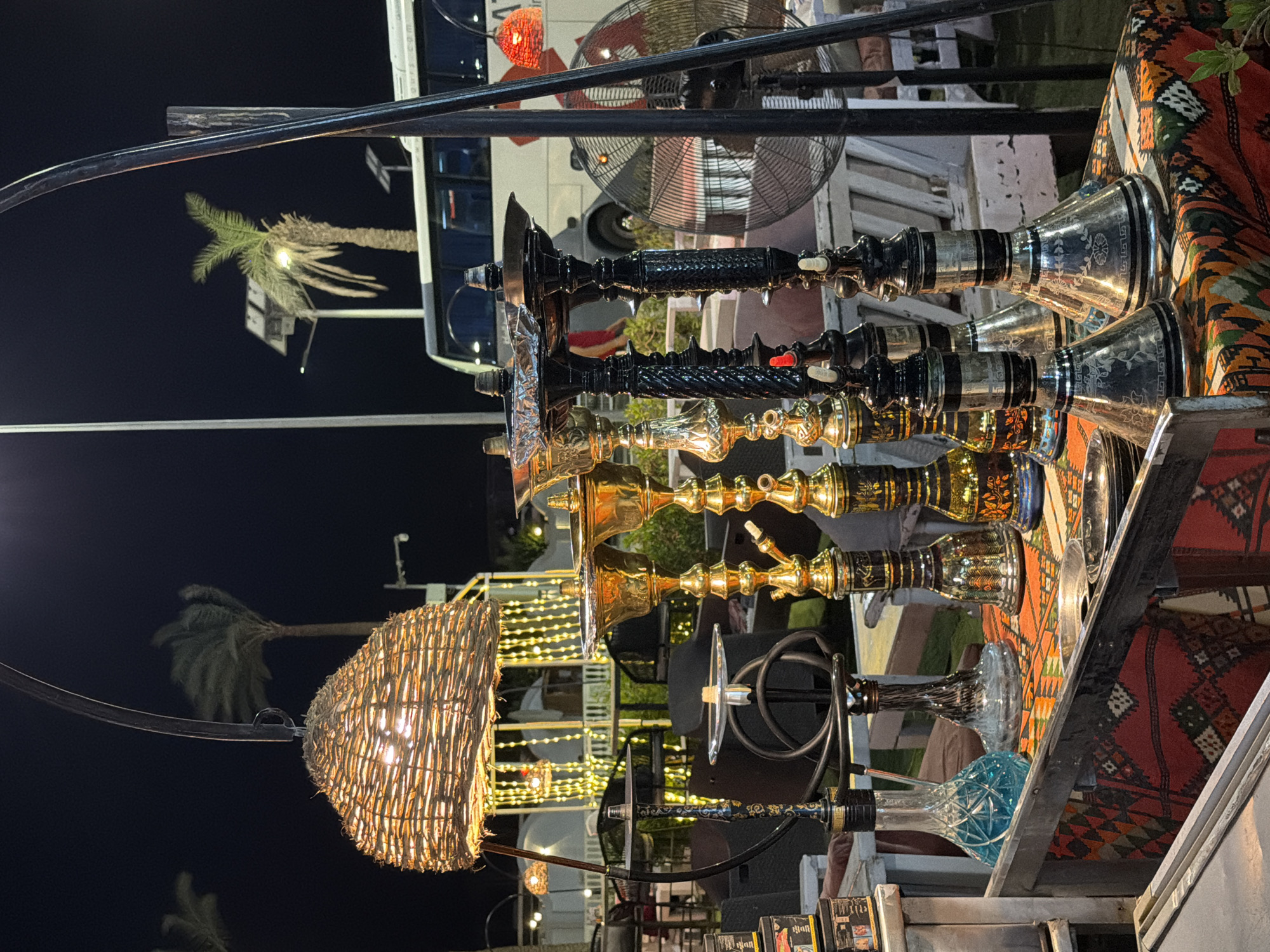 The image depicts an outdoor hookah lounge at night in Sharm El Sheikh, Egypt. In the foreground, a display of ornate hookahs of varying sizes and designs is set up on a table covered with a colorful, patterned cloth. The hookahs are made of metal and glass, some featuring intricate carvings and metallic finishes in gold and black. One hookah has a blue liquid in its glass base. Behind the hookahs, more seating areas with tables and chairs are visible, illuminated by string lights and ambient lighting. Palm trees and a street lamp with a solar panel are silhouetted against the dark night sky. A large tour bus is parked in the background, and a large industrial fan is positioned to the right of the hookahs. The overall scene suggests a relaxed evening setting.