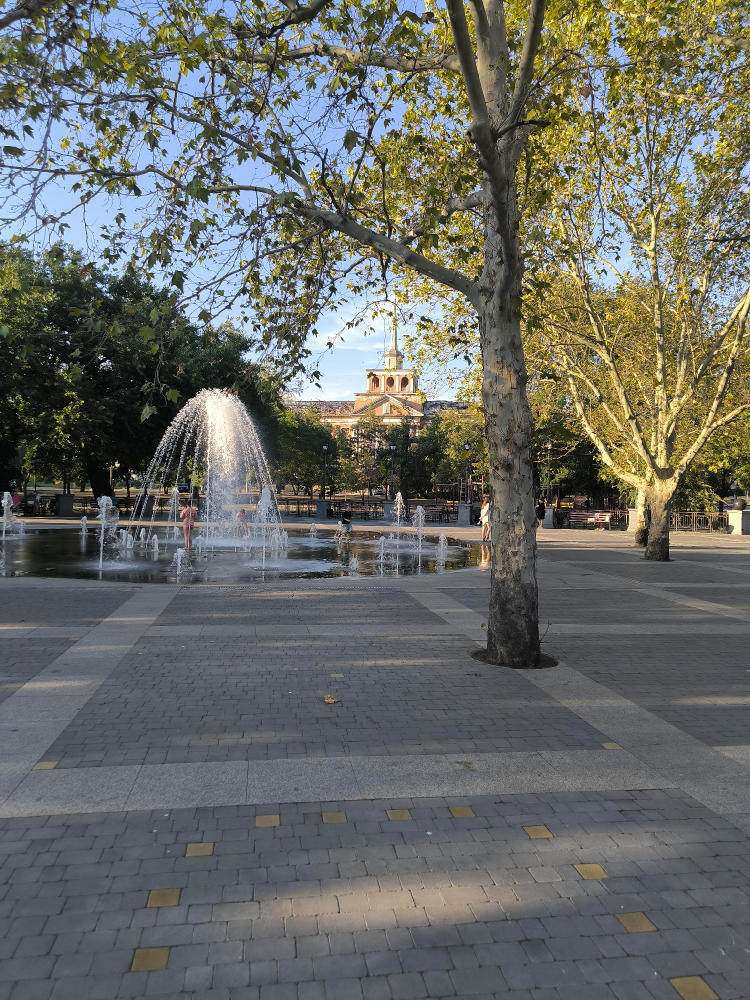 The image shows a public square with a fountain and trees. The fountain has multiple jets spraying water. There are several people, including children, playing in the fountain. Trees with yellowing leaves are prominent in the foreground and background, indicating autumn. A building with a distinctive domed tower is visible behind the trees. The ground is paved with grey and yellow square tiles. The overall scene depicts a recreational area within a city. This location is the Fountain Square (Skver Tsentralnyi) in Mykolaiv, Ukraine.