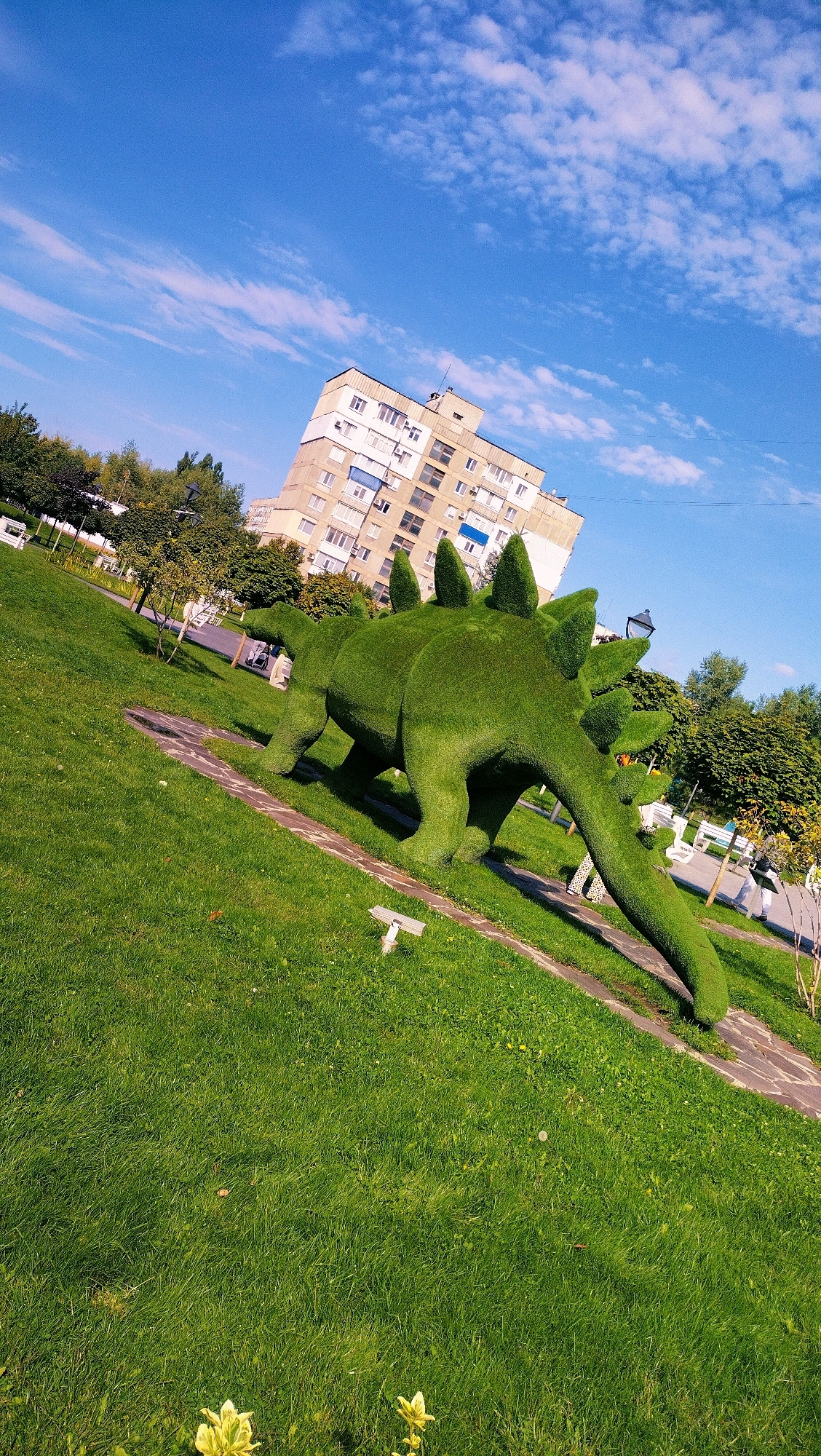 A green, topiary sculpture of a Stegosaurus dinosaur stands on a grassy lawn in a park in Horishni Plavni, Ukraine. The sculpture is the primary focus, with its distinct plates and spiked tail clearly visible. The park features well-maintained grass, a pathway, and trees. In the background, a multi-story apartment building with a light-colored facade and numerous windows is visible under a bright blue sky with scattered white clouds. The image appears to be taken during daylight hours on a clear day.