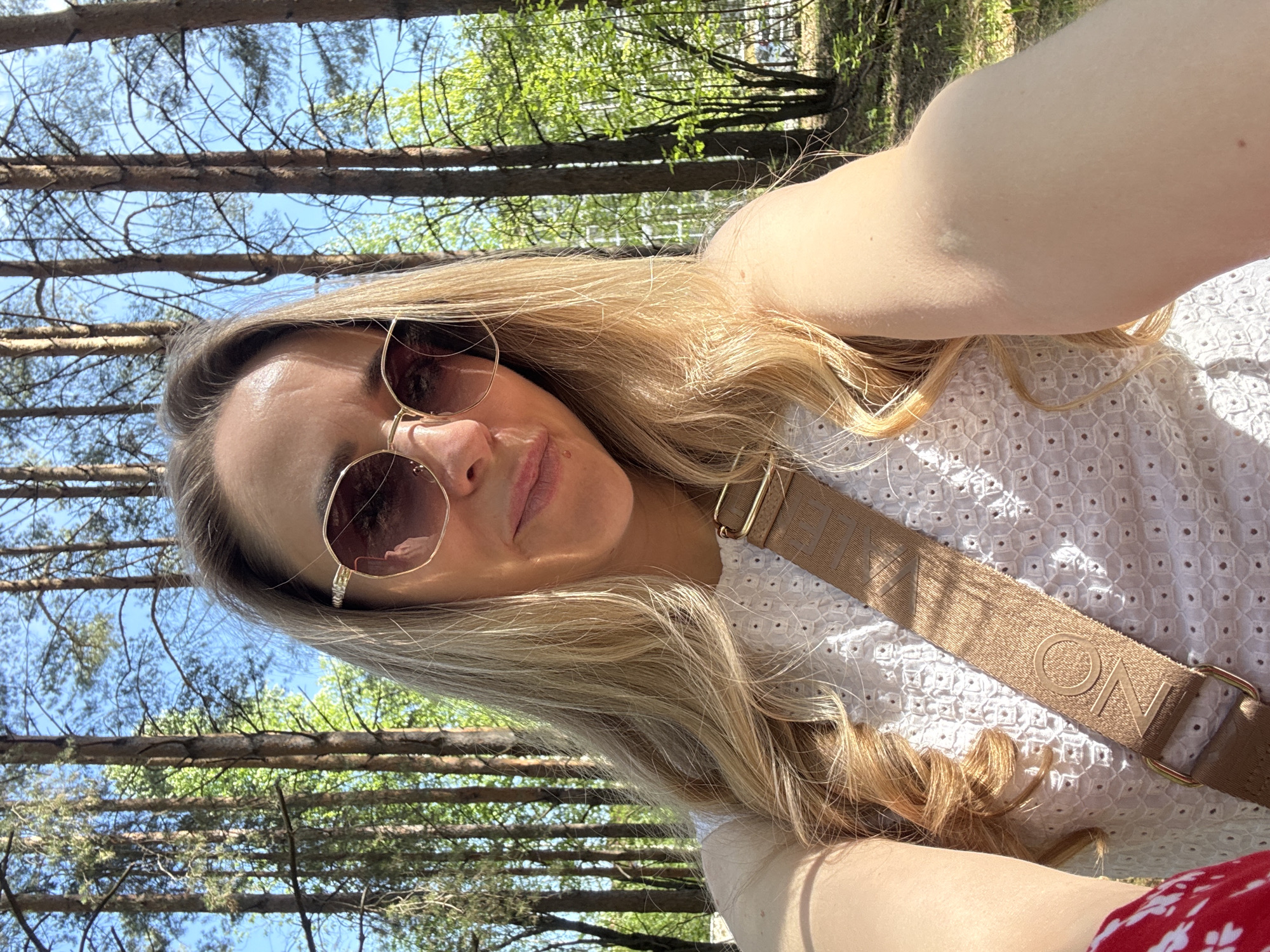 A woman is taking a selfie outdoors in a wooded area. She is wearing sunglasses and a white shirt with a brown strap over her shoulder. Trees and blue sky are visible in the background. No specific landmarks indicate Chaikovsky, Russia.