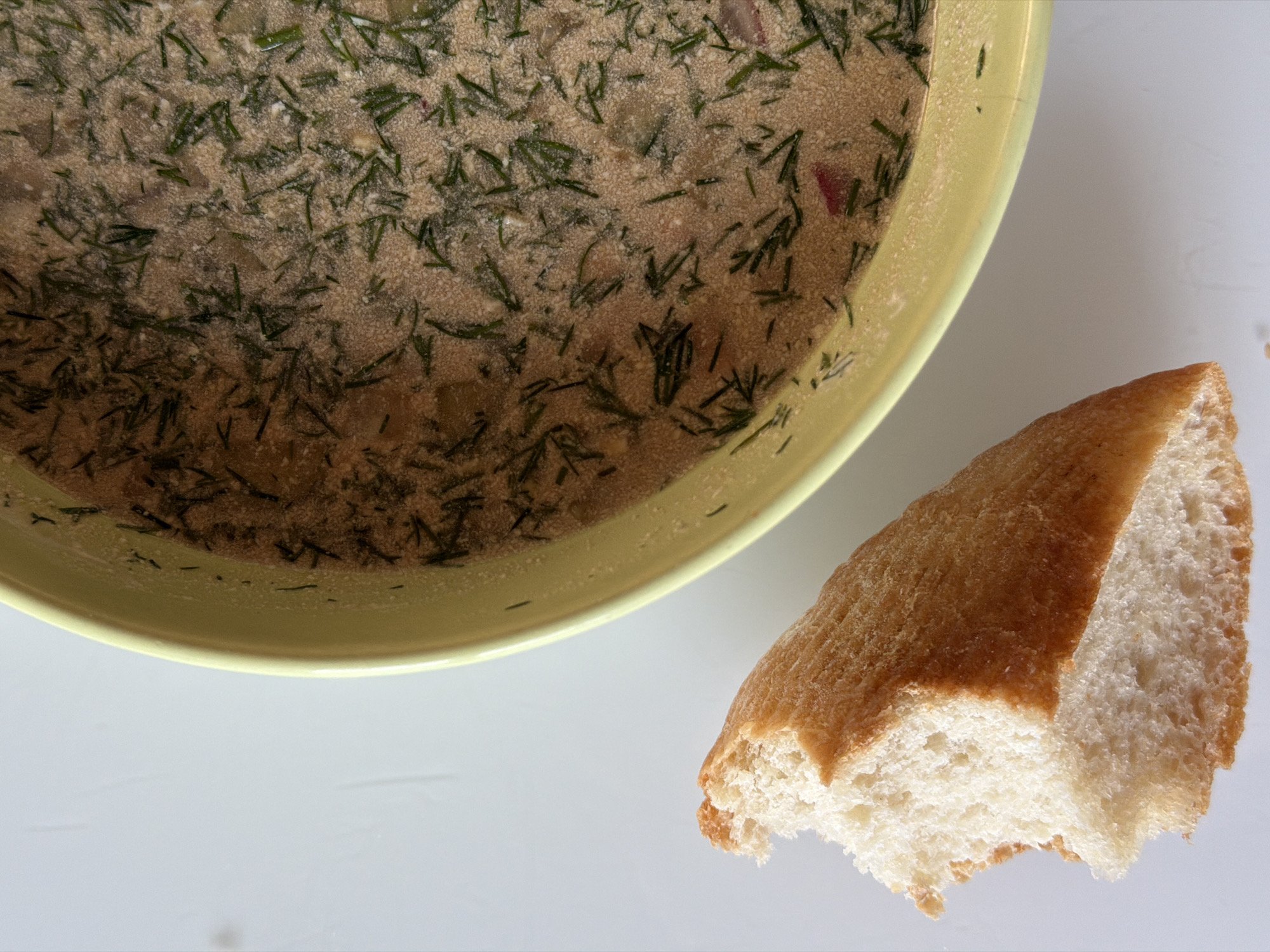 A close-up, overhead shot depicts a pale yellow bowl filled with soup, alongside a piece of bread on a white surface. The soup is speckled with green herbs, likely dill, and contains visible pieces of other ingredients. The bread has a golden-brown crust and a white, porous interior. The surface appears to be a white table or countertop. There is no information about the location in Chaikovsky, Russia, or any details regarding people, actions, or interactions visible in this image.