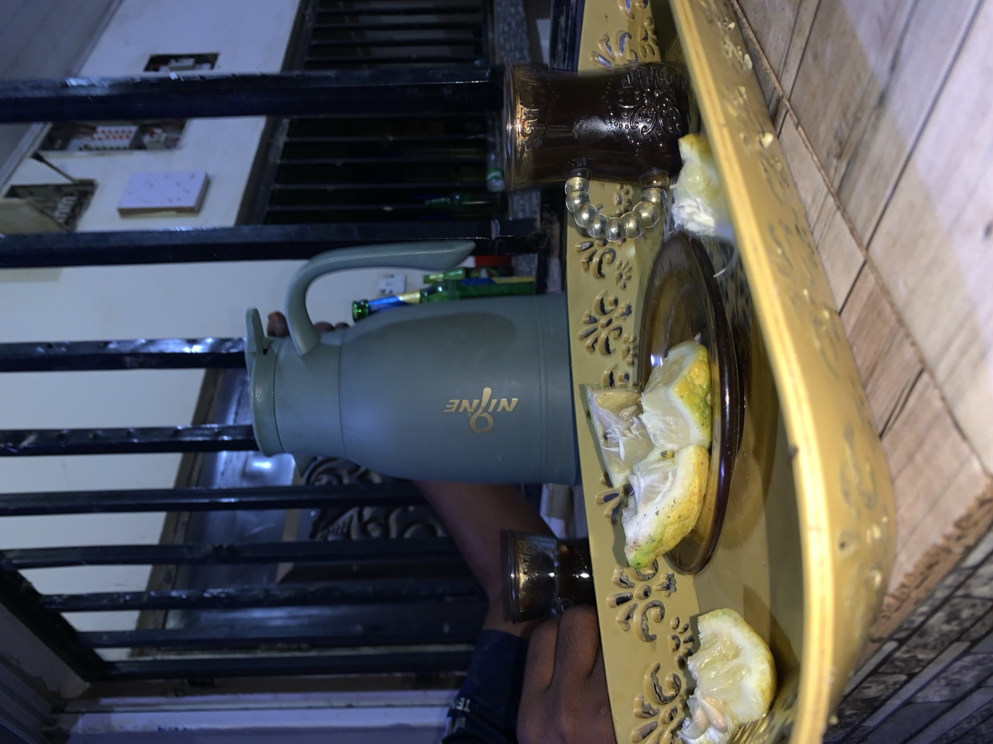 The image depicts a person, possibly in New Stadium, Nigeria, holding a tray with a pitcher labeled "Nine" and slices of lemon. There is a decorative cup on the tray as well. The setting appears to be indoors near a staircase with metal railings.