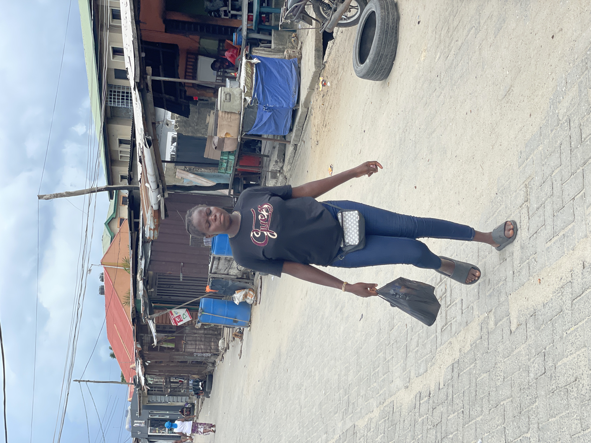A woman walks on a paved street in what appears to be a densely populated urban area, possibly a market or commercial district. She is wearing a black t-shirt with the word "Guess" printed on it, blue jeans, and gray sandals. She carries a black plastic bag in her right hand. A small, patterned crossbody bag hangs from her left shoulder. A spare tire leans against a wall in the background. The buildings along the street have corrugated metal siding and appear to be simple structures. There are various objects and structures visible, including blue barrels, signs, and other items. The sky is overcast.