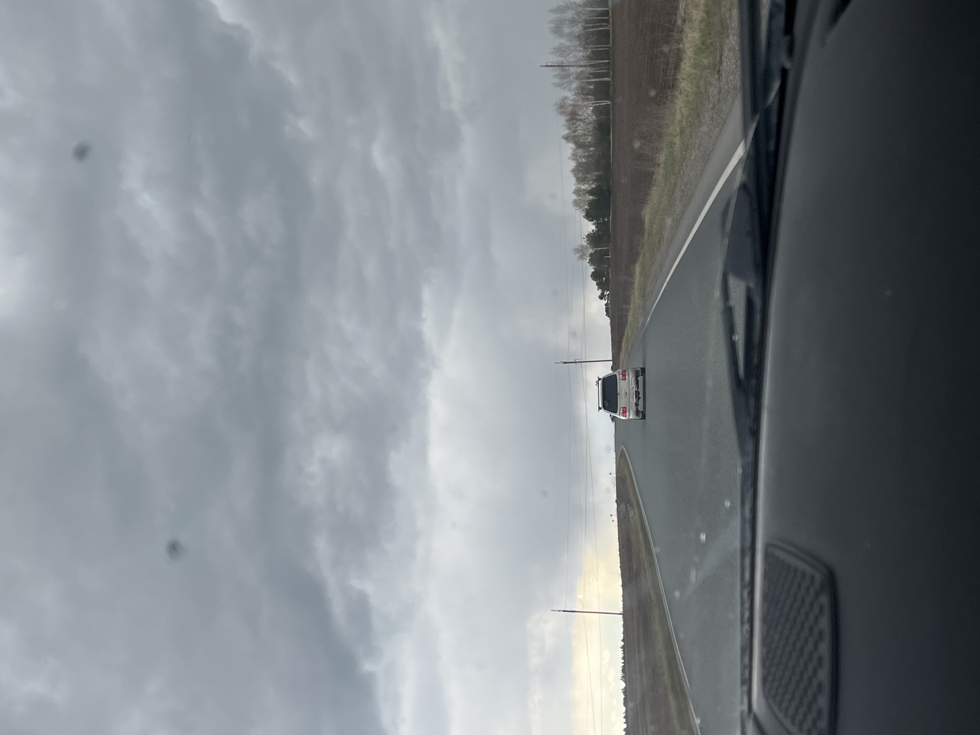 The image captures a perspective from inside a moving vehicle, looking forward down a rural road under an overcast sky.
In the immediate foreground, a dark grey or black section of the car's interior, possibly a door frame or dashboard, occupies the right side of the frame, indicating the viewpoint is from within the vehicle.
A two-lane paved road, appearing wet, stretches into the distance. White solid lines mark the road's edge on the right. Further down the road, a white pickup truck or SUV is visible, driving away from the viewer. Utility poles and power lines run parallel to the road on both sides, disappearing into the distance.
On either side of the road, agricultural fields extend. The field on the left appears to be freshly tilled earth, while the one on the right shows patches of grass and earth. A line of trees borders these fields in the mid-distance. Some trees are bare deciduous, possibly birches, while others retain their foliage.
The sky is heavily overcast with multiple shades of grey clouds, suggesting dull or rainy weather. A slight lightening of the sky is visible near the horizon, hinting at the sun's position or a break in the clouds. Several small, dark circular spots are visible against the sky, likely dust or imperfections on the camera lens or windshield.
The scene depicts a daytime journey through a rural landscape, characterized by very cloudy weather and a wet road, consistent with an ordinary drive in a countryside setting such as Ledukhi, Russia. No visible text is present in the image.