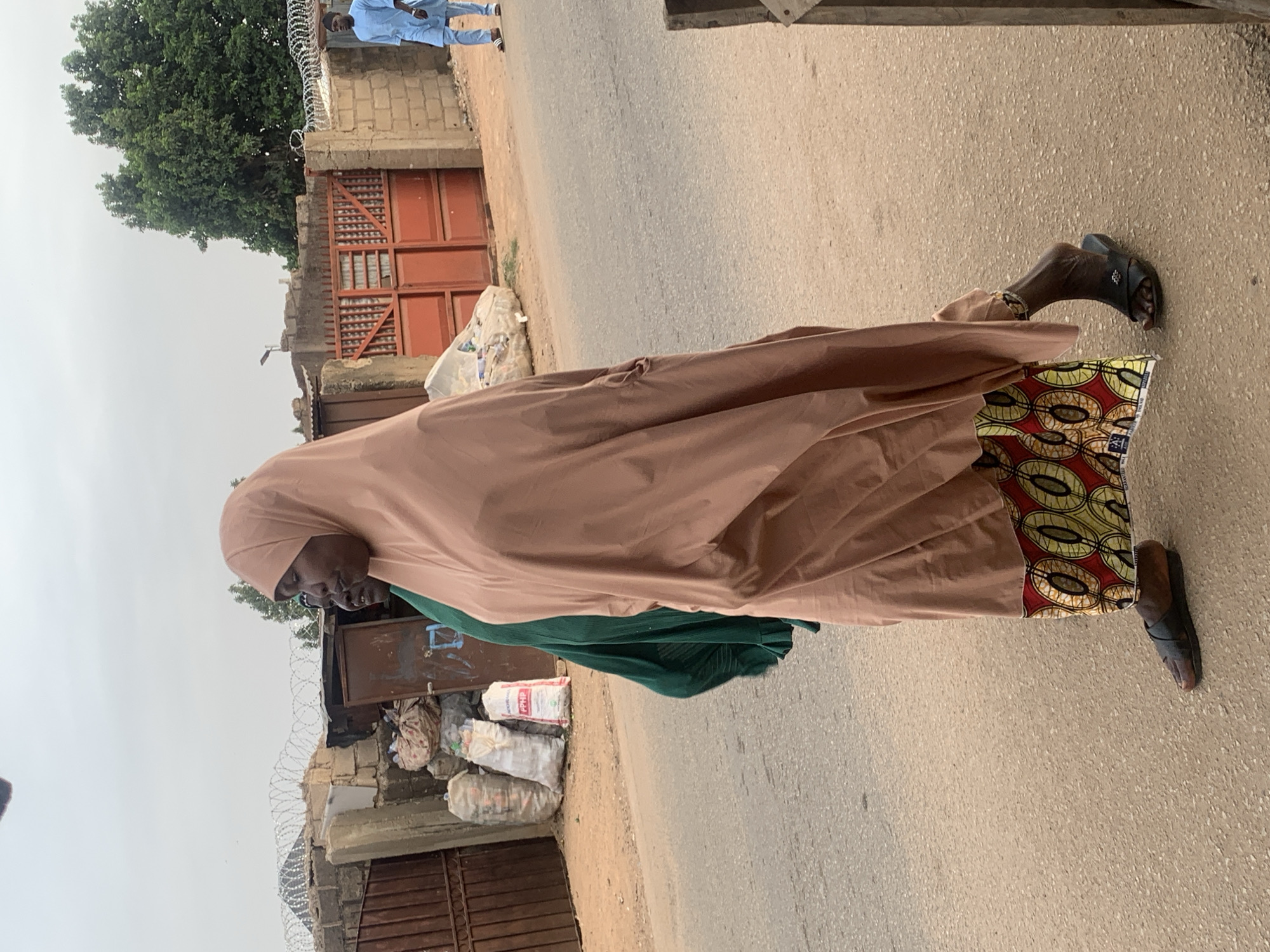 A woman wearing a tan hijab and a green and patterned dress stands on an asphalt road. She wears black sandals. Behind her are buildings with gates and barbed wire. Bags of what appears to be refuse are piled next to one building. Other people are seen in the background. The location is Jos, Nigeria.