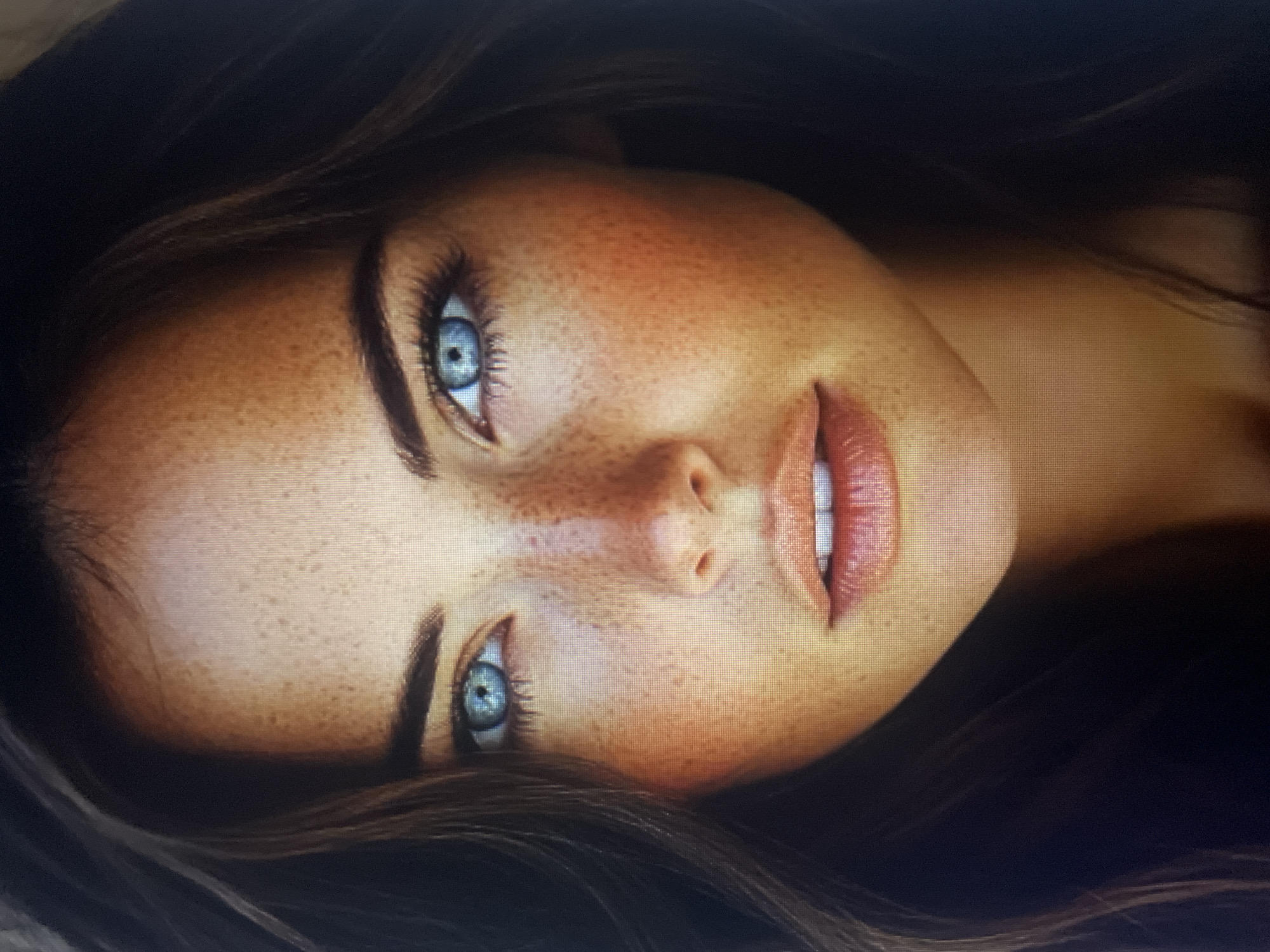 The image is a close-up portrait of a woman's face. She has fair skin with numerous freckles scattered across her cheeks, nose, and forehead. Her eyes are a striking bright blue with long eyelashes. Her lips are full and painted with a coral-colored lipstick. Her hair appears to be dark brown and is slightly blurred, framing her face. The lighting highlights her features, creating a warm glow on her skin. There is no discernible background or environmental context visible in the image. The location in Kołobrzeg, Poland, is not present in the image.