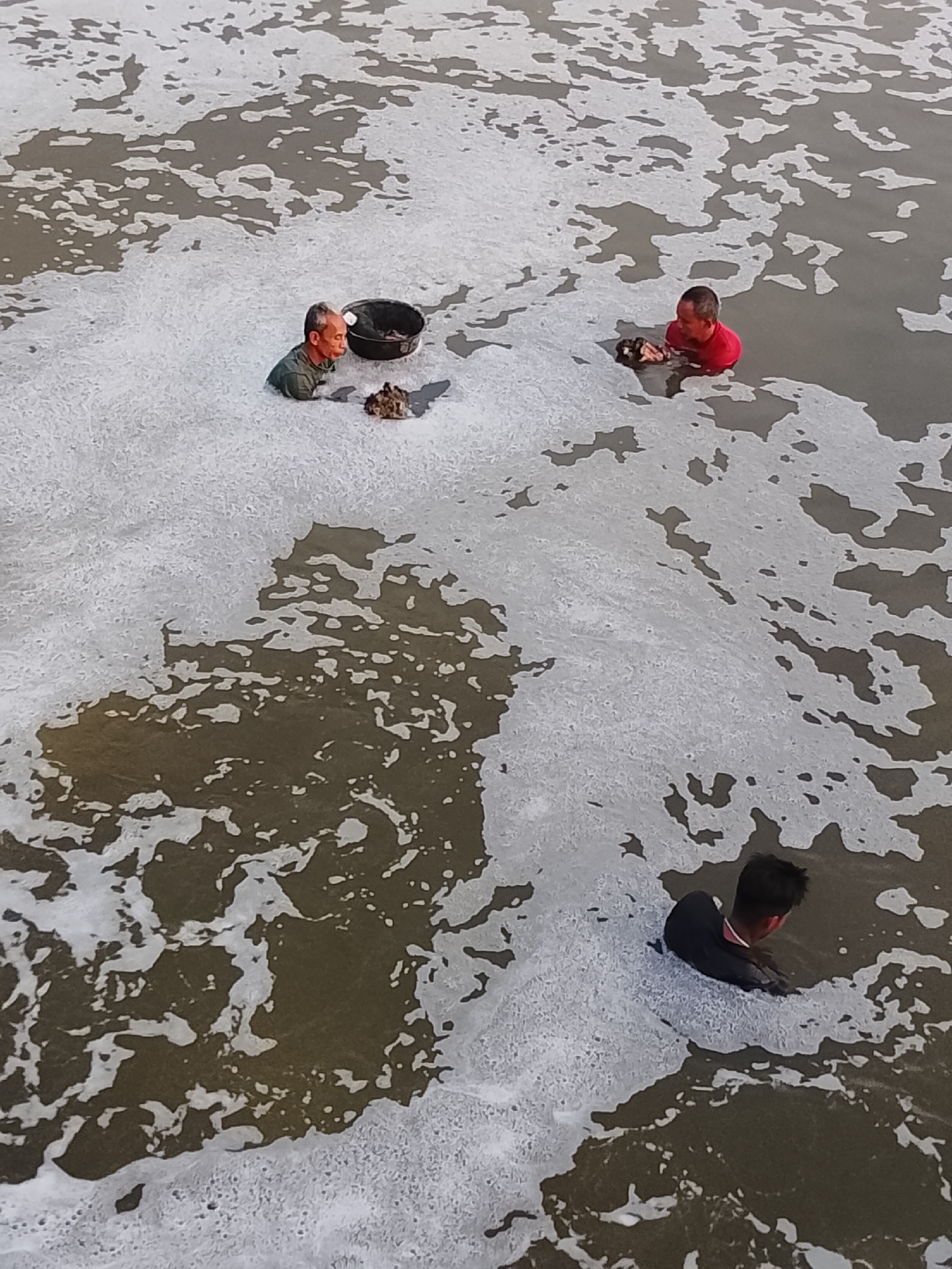 Three men are partially submerged in foamy, murky water. The man on the left wears a dark green t-shirt and is looking down at a black bucket and some debris. The man in the middle wears a red t-shirt and appears to be handling some debris in the water. The man on the right wears a black long-sleeved shirt and is facing away from the camera, with his head and shoulders visible above the water. The foamy water suggests that this scene may be taking place in an industrial area or near a source of pollution. The location is Lhokseumawe, Indonesia, a city known for its industrial activities, including an oil and gas processing plant, which could explain the presence of foamy water.