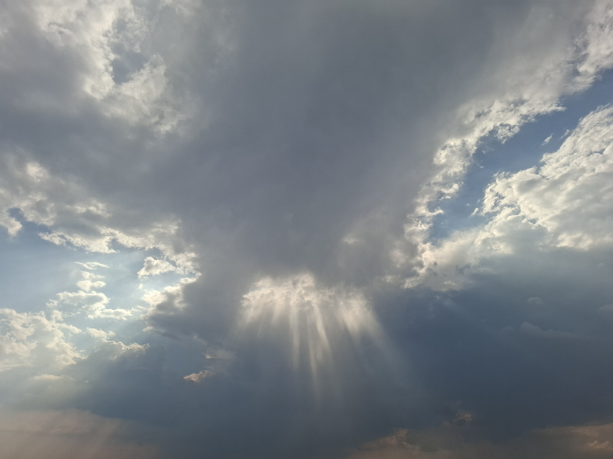 The image shows a dramatic sky scene with crepuscular rays piercing through dark, stormy clouds. The sun's rays are visible as bright shafts of light extending downwards from a break in the clouds. The sky is filled with a mixture of dark gray and white clouds, suggesting a dynamic weather system. There are no people, subjects, or objects present in the image, nor any actions or interactions taking place. The environment is solely the sky. The location of Topaly, Ukraine, is not depicted or inferable from the image itself.