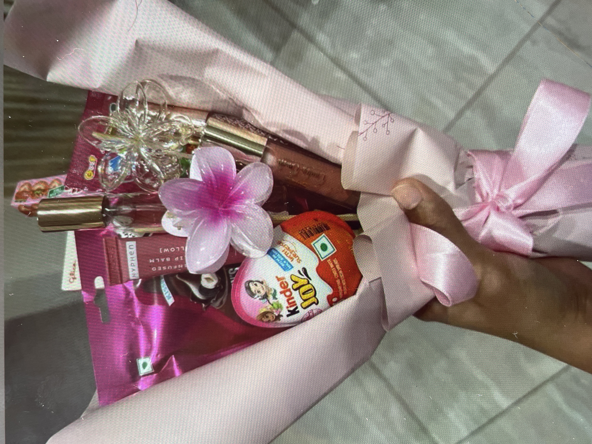 A bouquet of items is being held by a person. The bouquet is wrapped in pink paper and tied with a pink ribbon. The bouquet contains a Kinder Joy egg, a pink flower, a tube of lip gloss, a lip balm by "Hyphen", and a clear plastic flower. The location in Harmanli, Bulgaria cannot be determined from this image.