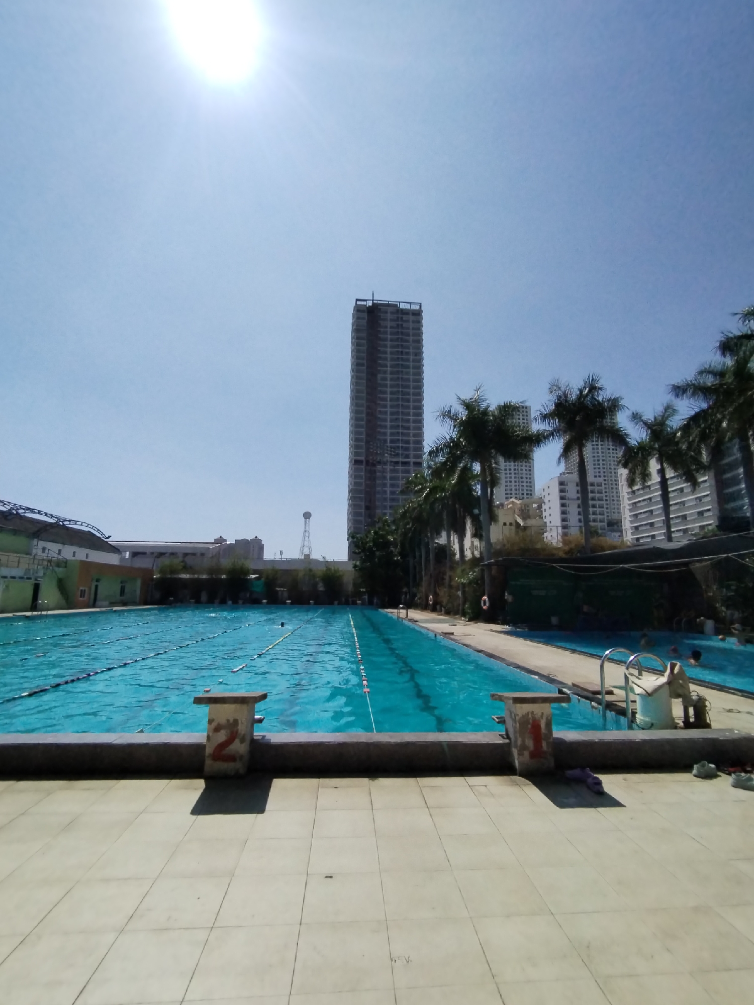 This outdoor scene depicts a swimming pool facility in Nha Trang Ward, Vietnam. The pool is filled with blue water and has lane markers stretching its length. There are starting blocks numbered "2" and "1" visible on the tiled pool deck in the foreground. Palm trees line the right side of the pool, with multiple buildings and a high-rise under construction in the background. The sky is clear and bright with a visible sun glare. A ladder and some equipment are visible on the edge of the second pool. Several people are in the water.