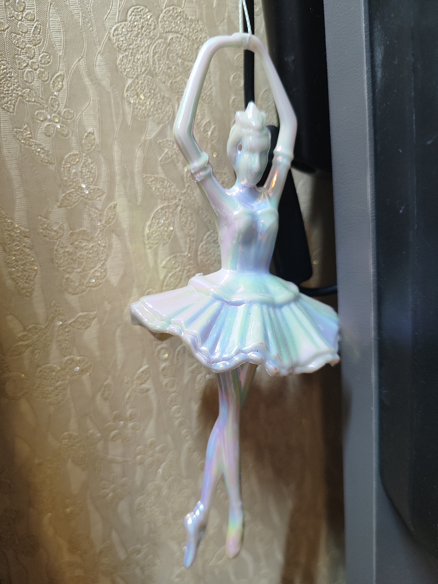 A close-up photograph features a white iridescent ballerina ornament hanging in front of a textured beige wallpaper and a black object. The ornament is detailed, with the ballerina's arms raised above her head, and she is wearing a tutu. Her feet are pointed as if in a ballet pose. The background includes a textured wallpaper with a floral pattern. The black object is out of focus, suggesting it is closer to the camera. The location is not discernible from the image.