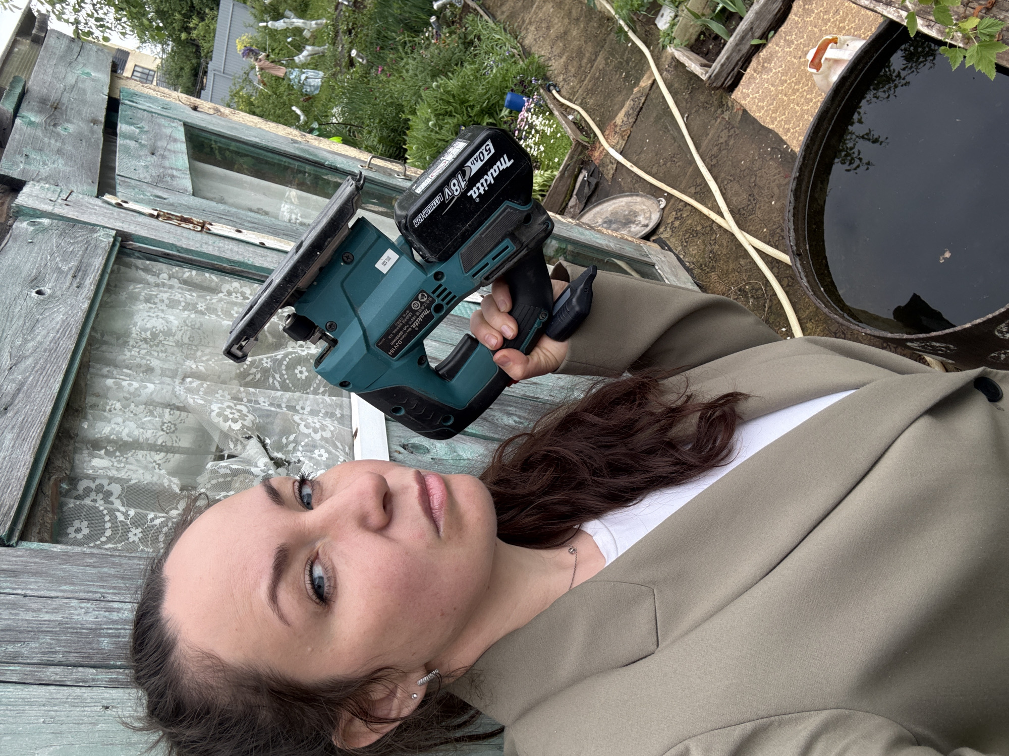 A woman lies on a weathered wooden surface with peeling green paint, holding a teal Makita jigsaw with a black battery pack. She is wearing a light brown blazer over a white shirt. Behind her is a garden area with various plants and a black barrel of water. The photo was taken in a garden setting, which could be inferred to be in Chaikovsky, Russia, based on the prompt. The location within Chaikovsky is unspecified, but the environment suggests a residential or backyard setting.