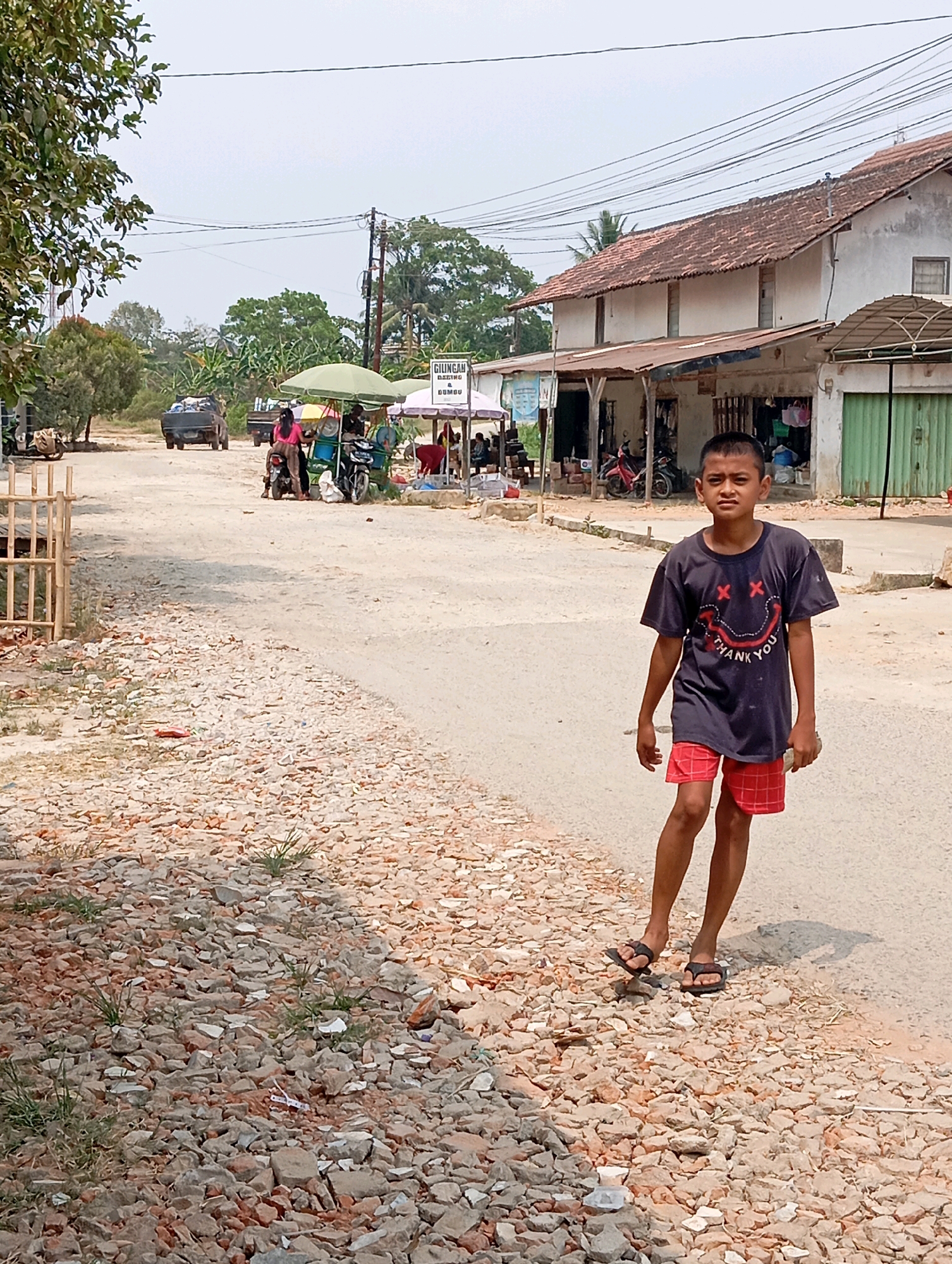 A young boy walks on a dirt road in front of a building. The boy is wearing a dark t-shirt with a smiley face design and red plaid shorts. He is also wearing black flip-flops. The building appears to be a shop or a series of shops. There are several motorcycles parked in front of the building, as well as some umbrellas and signs. The road is unpaved and covered with gravel and debris. In the background, there are trees and a clear sky. The scene appears to be in a rural or semi-urban area. The location is identified as Batang Hari Ogan, Indonesia.