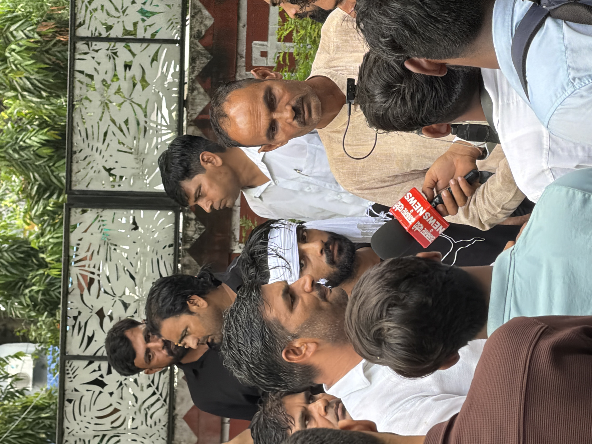 The image depicts a group of men gathered outdoors, possibly for a press conference or interview. The subjects are diverse in age and appearance, with several men in the foreground looking towards a camera or reporter. One man in a light-colored shirt has a microphone clipped to his collar and is holding another microphone. Another man in the foreground wears a white cloth headband and has a beard. The setting appears to be in New Delhi, India, with a textured wall and foliage visible in the background. The overall atmosphere seems serious, with the men appearing focused and perhaps concerned. The time of day is not clearly indicated, but the lighting suggests it could be daytime. Visible text on a red object held by one of the men reads "NEWS".