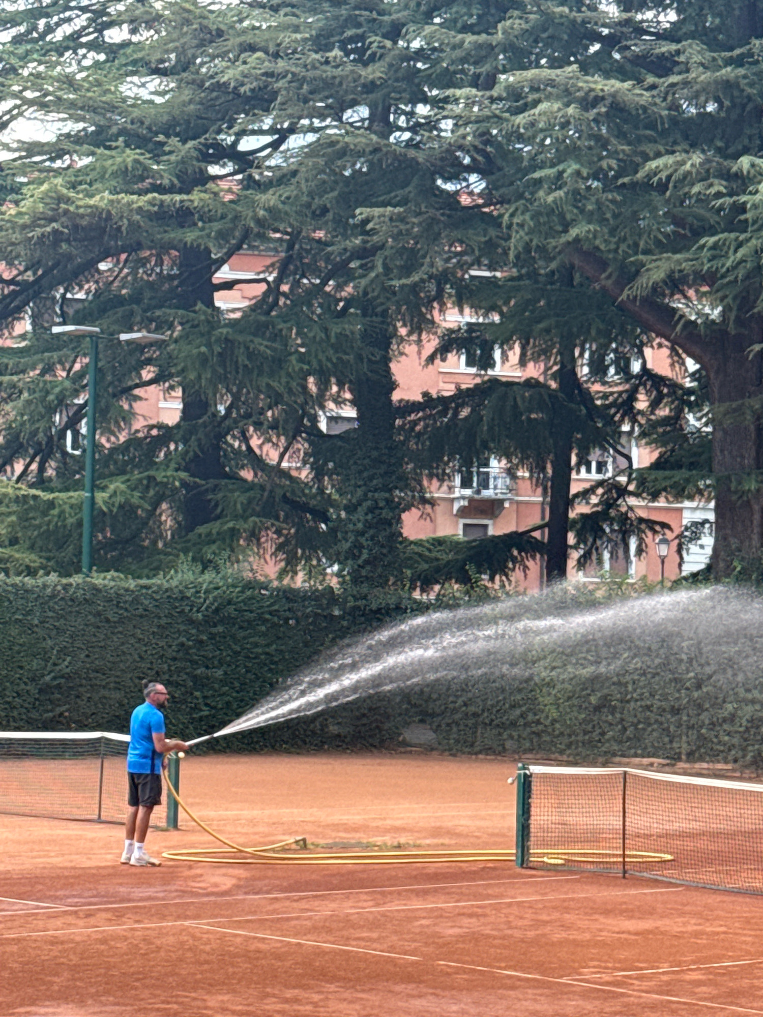 A man with a bun hairstyle, wearing a blue t-shirt and black shorts, stands on a clay tennis court in the city of Trento, Italy. He is holding a yellow hose, watering the court with a wide spray of water. The court has white lines and a net. In the background, a dense green hedge lines the court, behind which is a multi-story building with salmon-colored walls and white-framed windows. Large, mature trees with dark green foliage overhang the scene. The lighting suggests an overcast day.