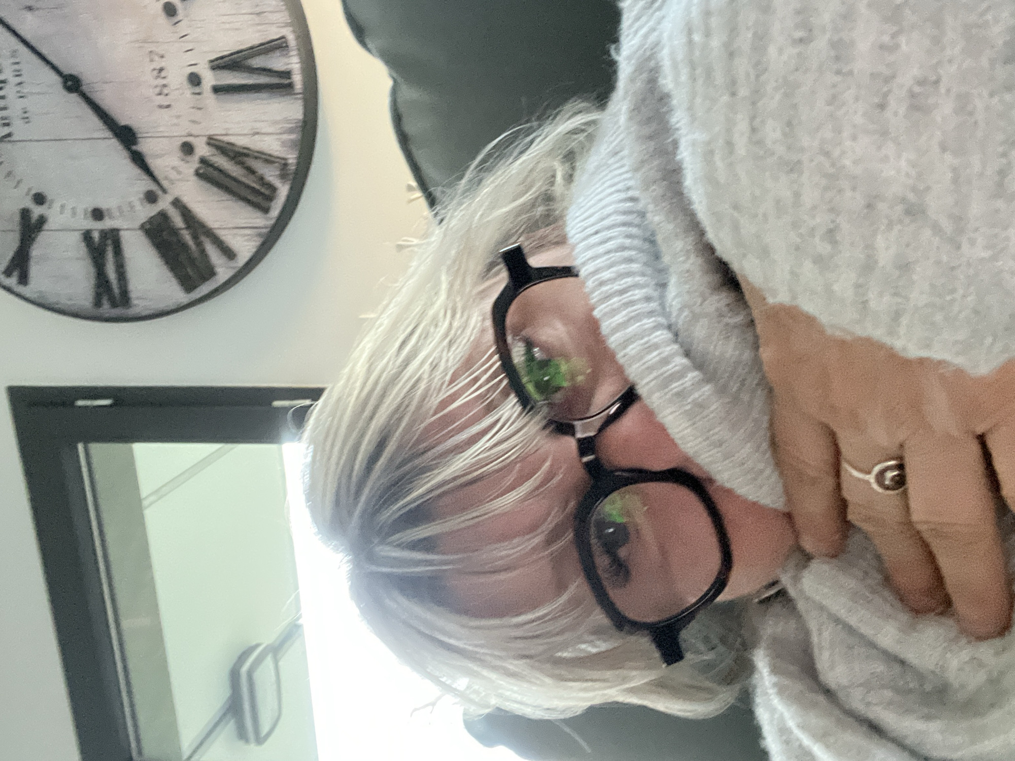 The image is a close-up, slightly angled view of a woman with short, blonde hair wearing black-framed glasses and a light grey sweater. Part of her face is visible, including her eyes behind the glasses, nose, and mouth, along with her hand with a ring on one of the fingers. In the background, there is a clock hanging on the wall showing the time at approximately 10:50, and a black-framed mirror reflecting light. The location of the photo is unknown; therefore, there is no Chambœuf, France connection possible.
