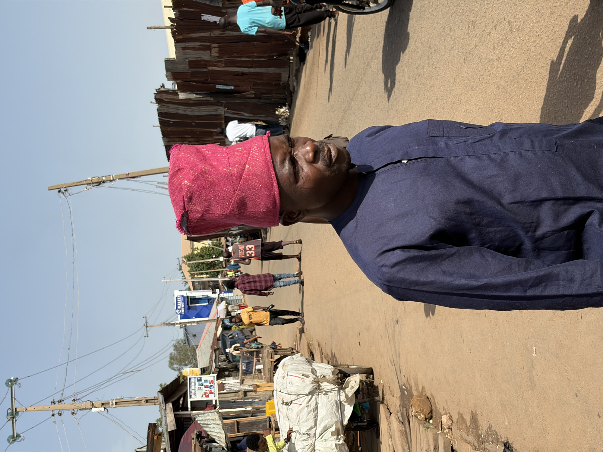 The image shows a man wearing a dark blue shirt and a red cap, standing on a street in Gura Top, Nigeria. In the background, there are pedestrians, a bicycle, shops with corrugated metal walls, telephone poles, and a cart with a large white bundle. The street is unpaved.