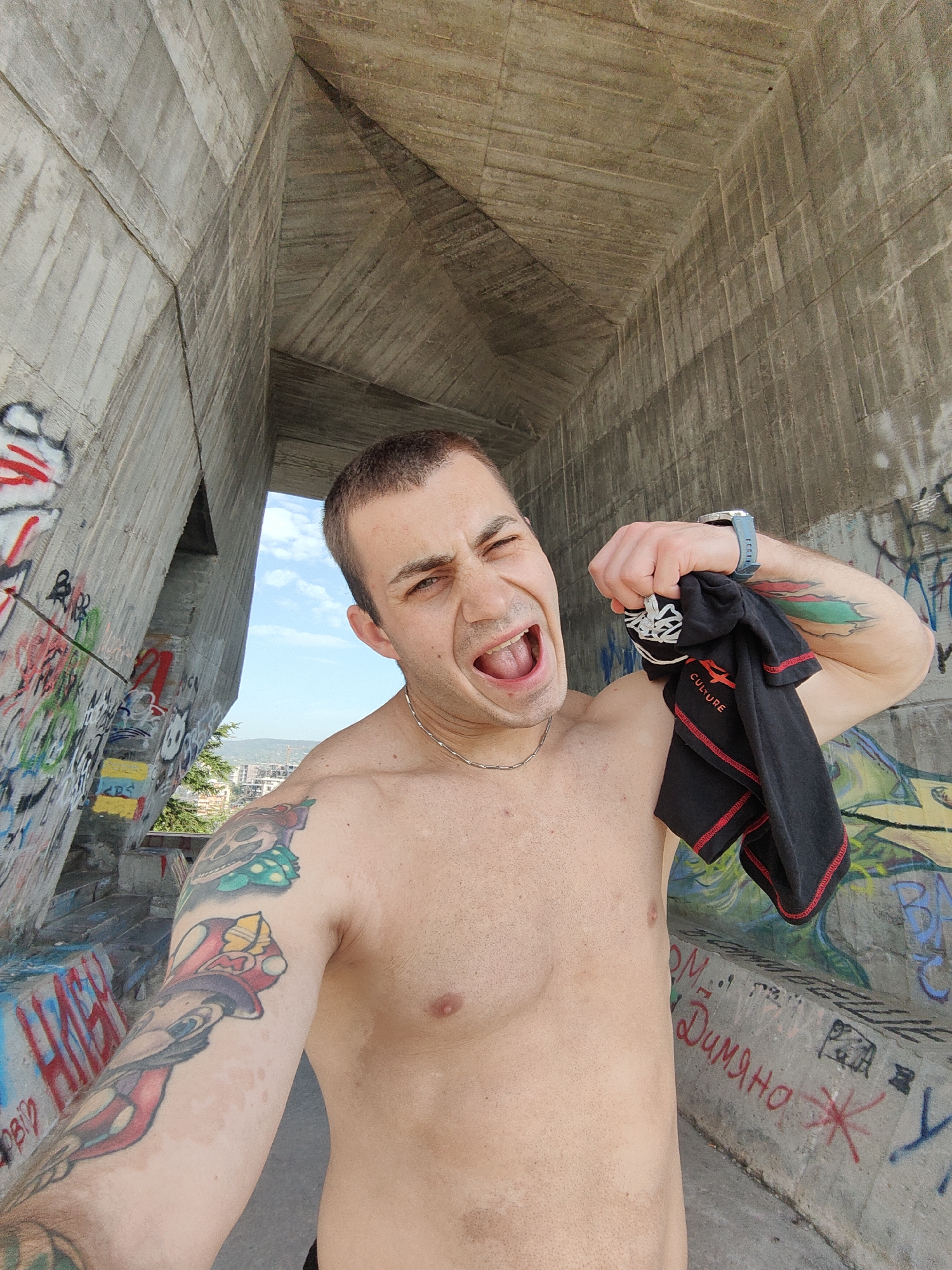 A shirtless man with tattoos on his right arm is taking a selfie. He has a short haircut, a silver chain around his neck, and a blue wristwatch on his left wrist. He is holding a black T-shirt with red trim in his left hand and making a wide-mouthed expression.
He is standing within a concrete structure covered in graffiti. In the background is a view of the city of Varna, Bulgaria, partially obscured by the structure. The sky is blue.