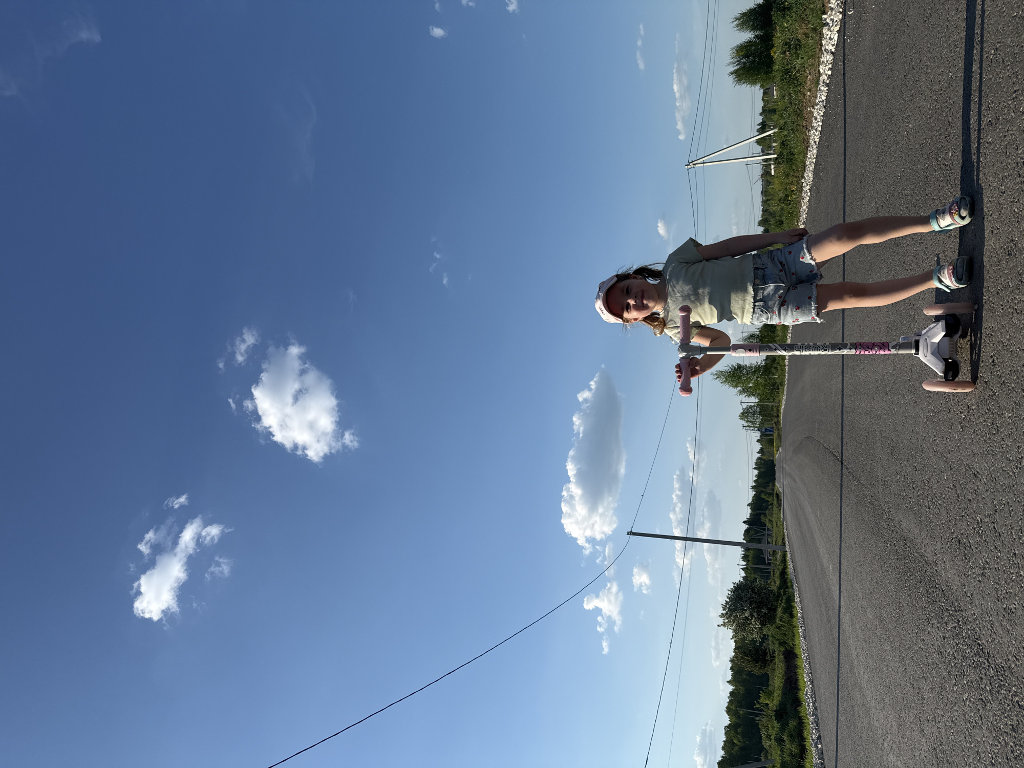 A young girl stands on a scooter on a paved road under a clear blue sky with scattered clouds. She is wearing a light-colored t-shirt, jean shorts with strawberry patterns, a white baseball cap, and sandals. The scooter is white and pink. The road is asphalt, and vegetation lines both sides. Electrical poles and lines are visible. The location is Izhevsk, Russia.