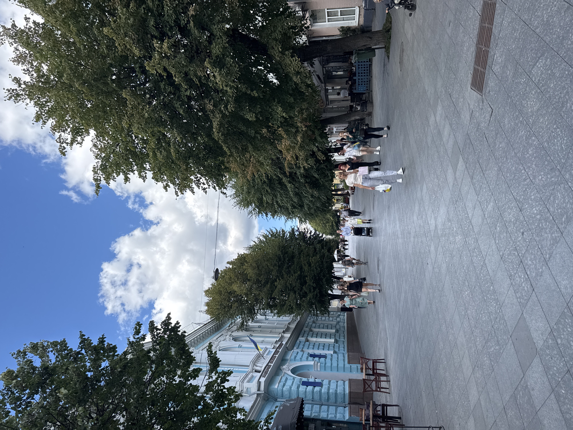 The image captures a street scene in an urban environment, likely a pedestrianized area, on a bright day with scattered clouds in a blue sky. On the left side of the image, a light blue building with ornate white trim and arched windows dominates the foreground. A Ukrainian flag is displayed on the building. Sidewalk cafes with tables and chairs are set up in front of some establishments. A tree-lined walkway stretches into the distance, with mature trees providing shade. Several people are walking along the pedestrian path, some are carrying shopping bags, one person is riding a bicycle, and another is pushing a stroller. The ground is paved with large gray tiles. The overall atmosphere suggests a pleasant day for a stroll or outdoor dining.