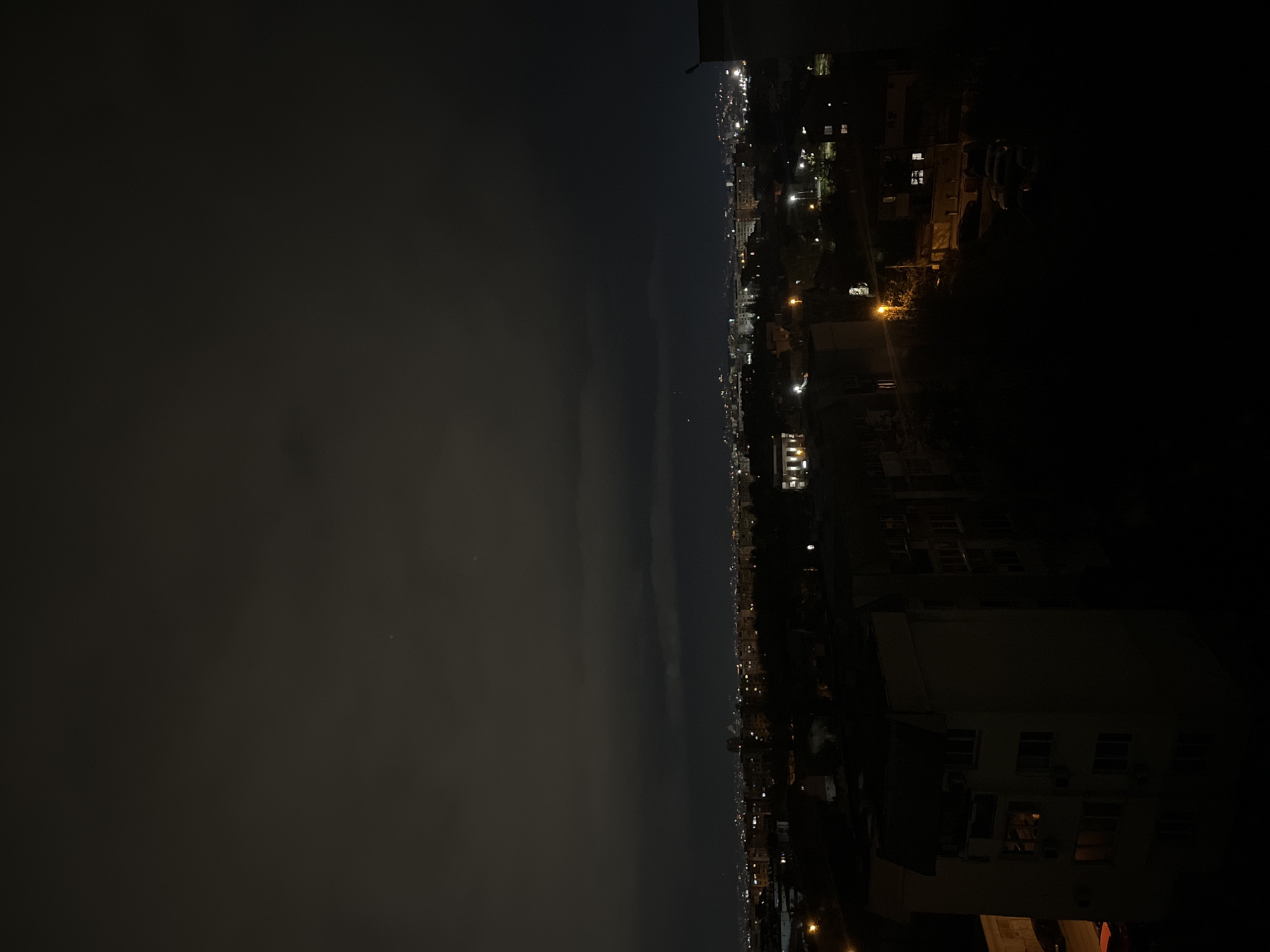 The image captures a wide, panoramic view of a city at night, likely Sofia, Bulgaria, from an elevated vantage point. The scene is bisected by a distinct horizon line, separating the dark night sky from the illuminated urban landscape.
The upper portion depicts a dark sky, which appears hazy or partially overcast, with subtle variations in cloud density. Faint, distant points of light, possibly stars or very high-altitude objects, are barely discernible within the atmospheric haze.
Below the horizon, the city stretches across the frame, a dense network of artificial lights. Numerous building lights, streetlights, and other urban illuminations create a vibrant glow, indicating active urban life. In the foreground and mid-ground, particularly on the right side of the image, the silhouettes of several buildings are visible, some with warmly lit windows. Streetlights cast pools of light, defining thoroughfares and residential areas. The city lights extend into the distance, gradually fading into the nocturnal haze near the horizon.
No individuals or specific activities are identifiable. The image conveys the tranquil yet energetic ambiance of a large city after dusk. The atmospheric conditions suggest a calm night, possibly with some light cloud cover or urban light pollution contributing to the sky's appearance. There is no visible text within the image.