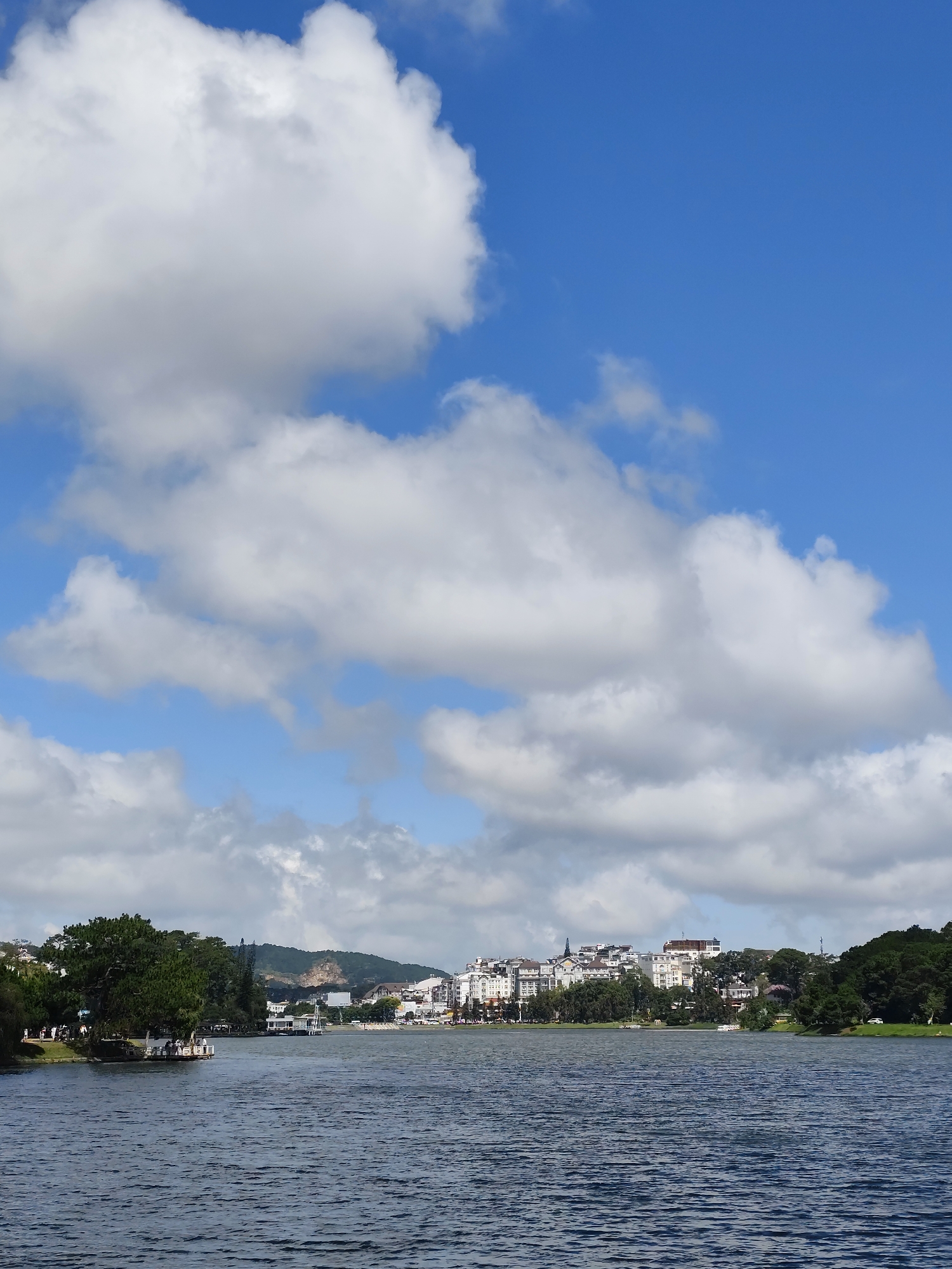 The image depicts a scenic view of Xuan Huong Lake in Da Lat, Vietnam, under a partly cloudy blue sky. The foreground is dominated by the dark blue, rippling water of the lake. Along the far shore, a line of buildings, primarily white and beige, forms a townscape that stretches across the horizon, punctuated by lush green trees and hills in the background. To the left, a verdant tree-covered area meets the water, with a wooden pier extending into the lake, occupied by several people and a boat. Further along the shore, more boats are visible on the water, and the town continues with its white-colored architecture. The sky above is a bright blue, with large, fluffy white clouds scattered across it, adding depth and texture to the scene. The overall impression is one of a tranquil, picturesque lakeside urban environment.