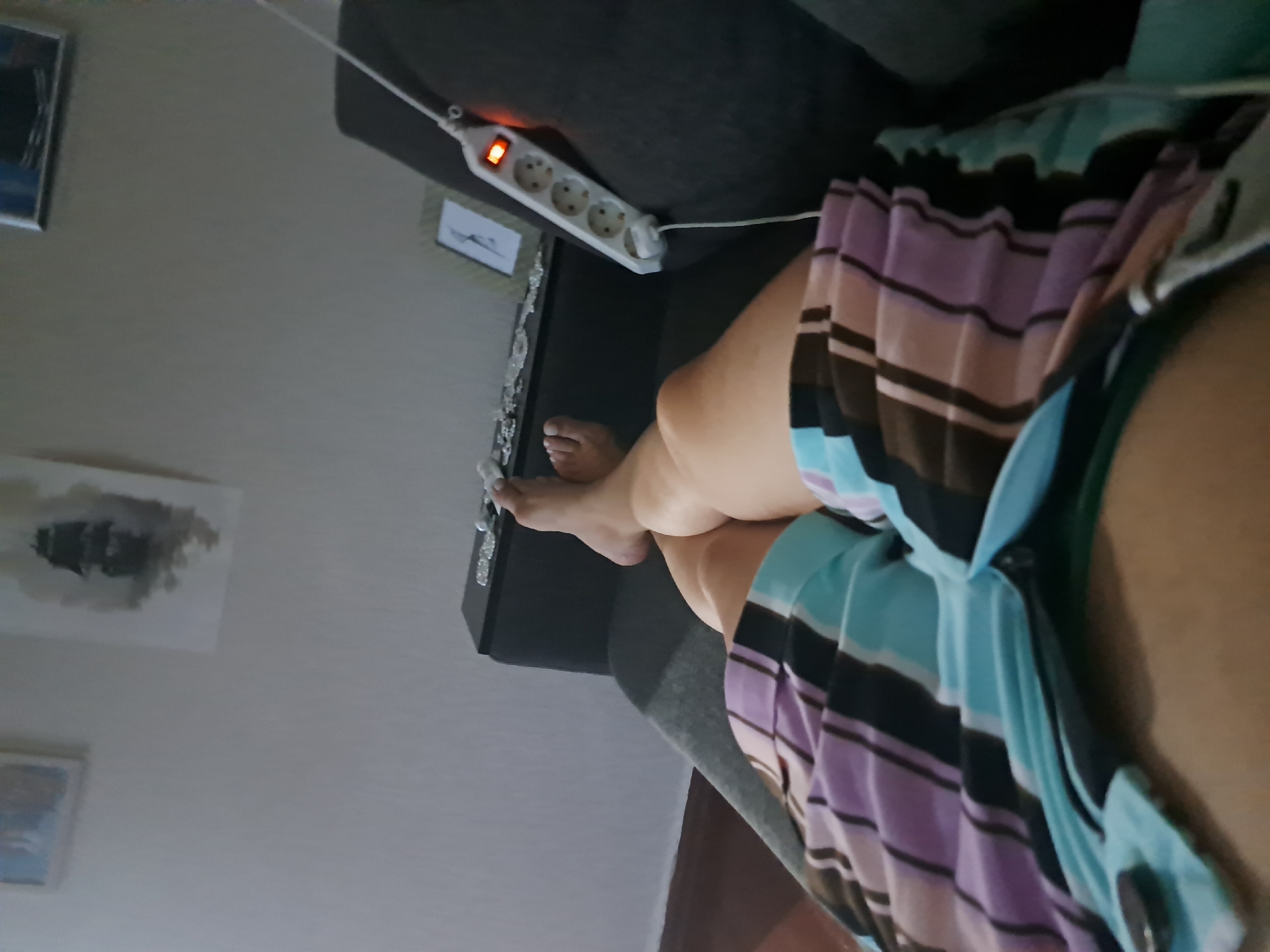 The image shows a person's legs and torso, seen from a low angle, resting on a gray couch. The person is wearing striped shorts in shades of purple, black, and light blue. The background shows a wall with framed artwork and a black furniture piece. To the right, a white power strip with an illuminated orange switch is plugged into an outlet. The scene appears to be indoors, likely a living room. No specific location in Leningradskaya, Russia can be determined from the image.