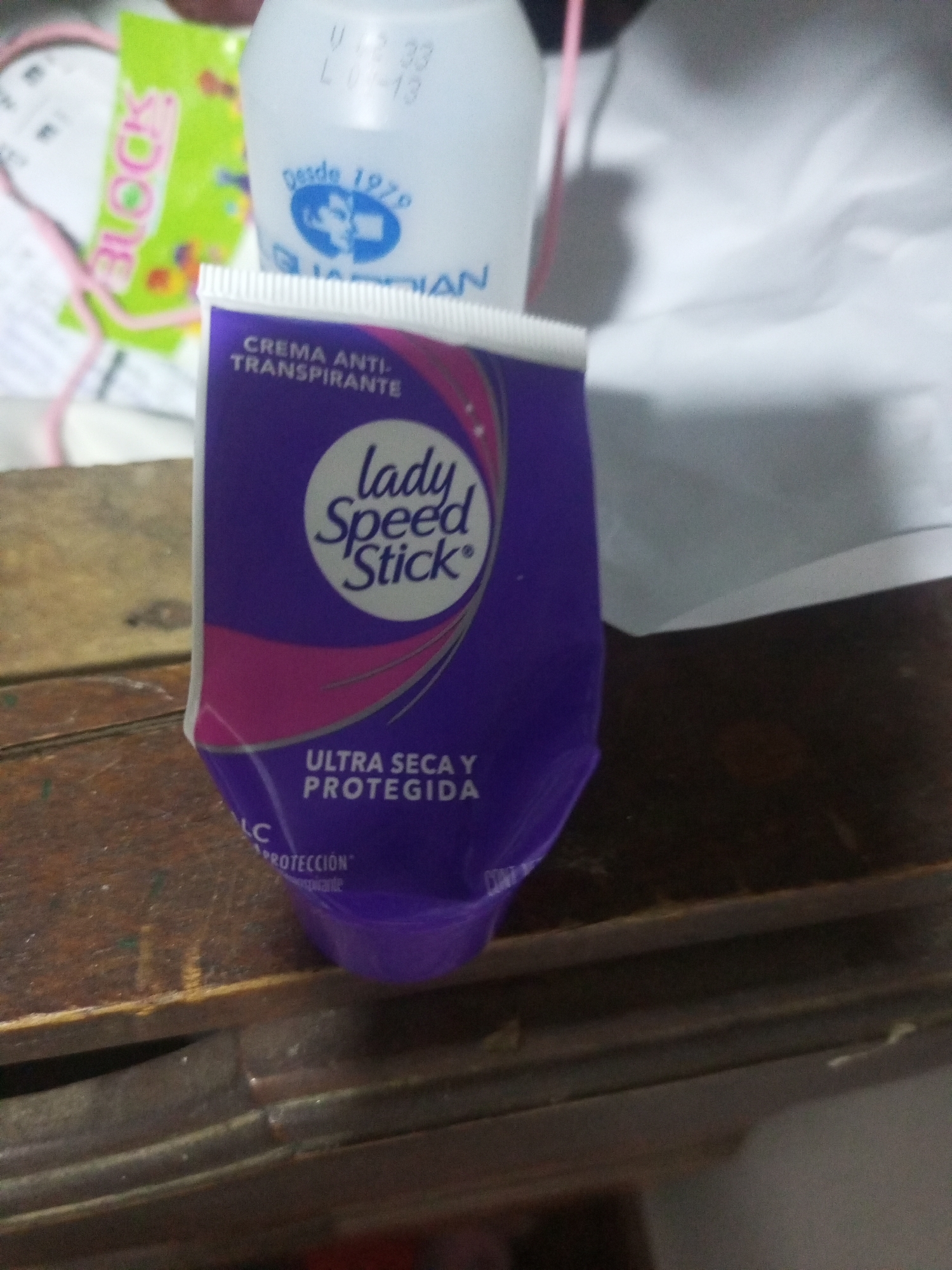 A purple and pink tube of Lady Speed Stick deodorant is centered in the foreground of the image, resting on a wooden surface. The tube has white text on it that reads "CREMA ANTI-TRANSPIRANTE ULTRA SECA Y PROTEGIDA." A white bottle with a blue logo that reads "Desde 1979" is visible behind the deodorant tube. A green package with the word "BLOCK" printed in pink letters is to the left of the deodorant tube. The background is out of focus, showing a white sheet or fabric. The overall scene appears to be indoors, with the wooden surface suggesting a table or shelf. There are no people visible in the image. The provided location of La Tagua, Venezuela, is not visually discernible from the image itself.