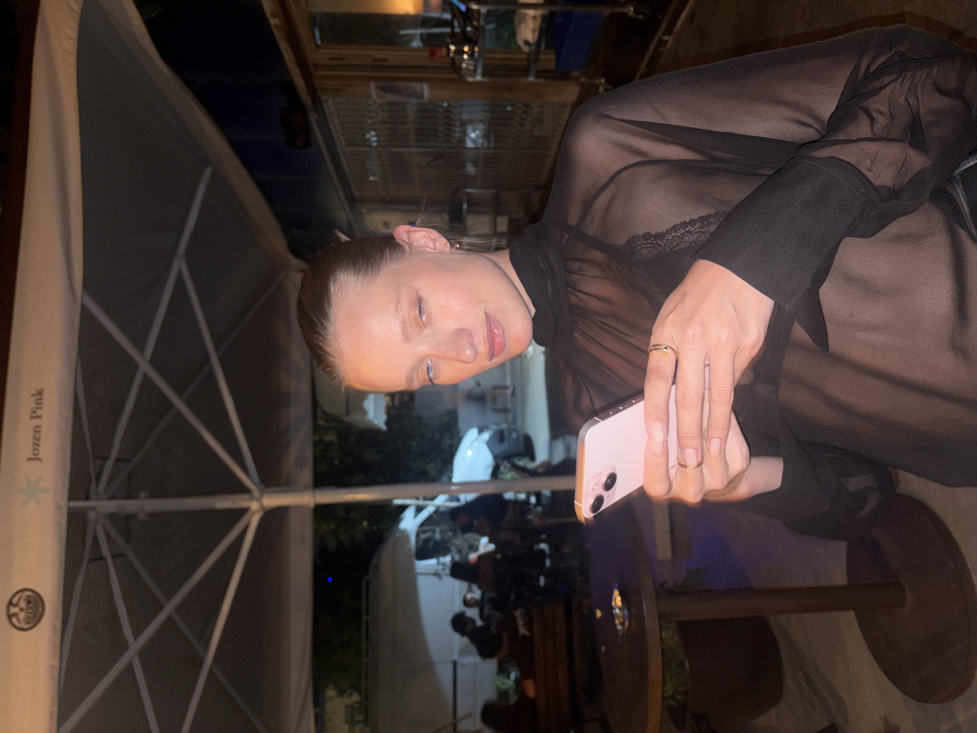 A young woman with light skin, blonde hair pulled back, and blue eyes is standing outdoors at night, holding a pink smartphone in her hands. She is wearing a sheer black top with a high neckline and lace detailing on the chest. She is looking down at her phone, with a slight smile. In the background, there are people sitting at tables under an umbrella, and a white van parked. The scene appears to be a cafe or restaurant in Tel Aviv, Israel. The overall atmosphere is casual and social.