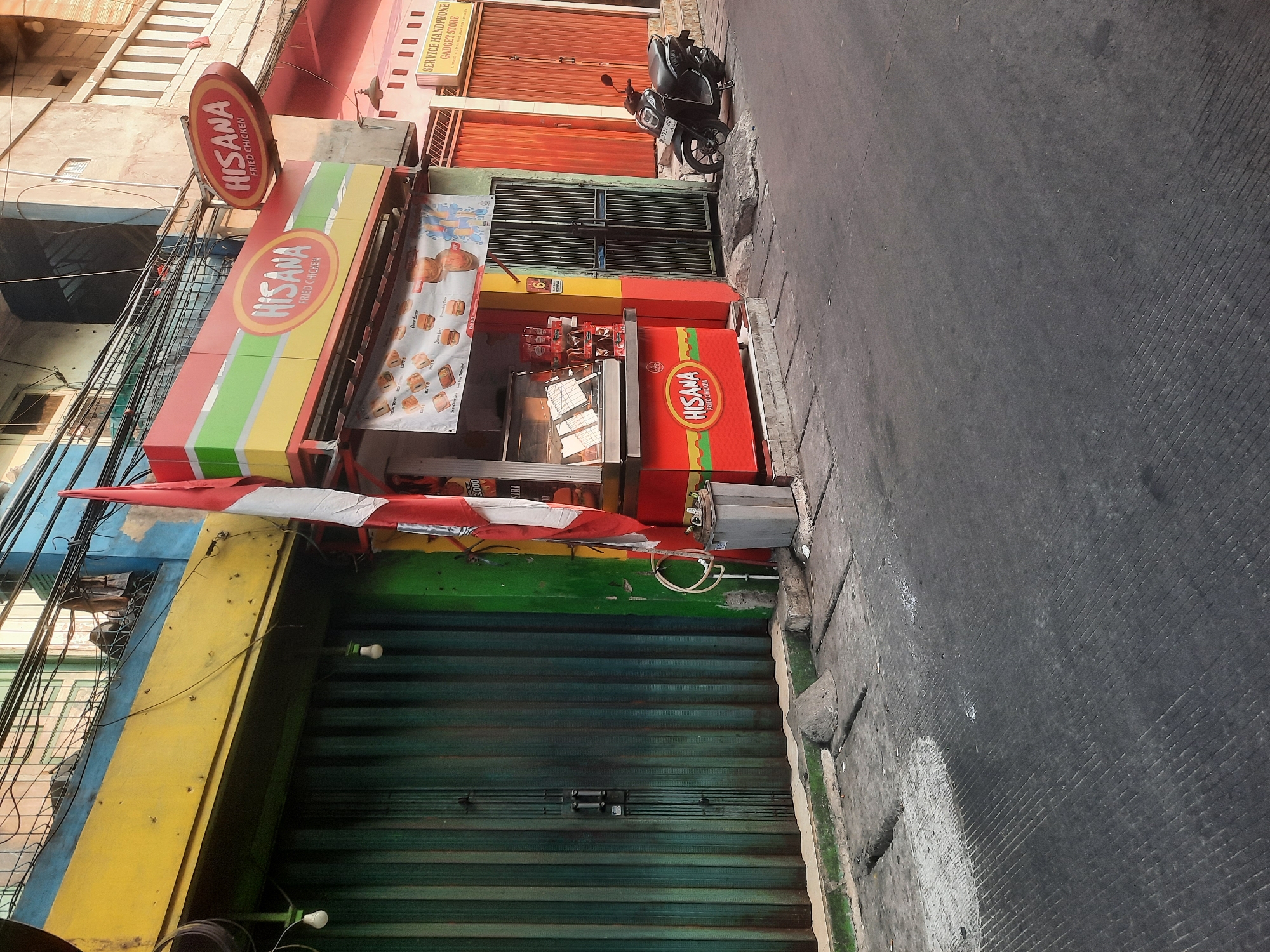 The image shows a street scene in North Jakarta, Indonesia. In the foreground, a dark asphalt road with diagonal grooved markings stretches into the distance. On the left side of the road, there is a green metal rolling shutter door that is partially closed, with a light bulb hanging in front of it. Adjacent to the shutter is a yellow-painted section of a building.
In the center of the image, a bright red food stall with the "HISANA FRIED CHICKEN" logo is prominently displayed. The stall has a display case with food items and a menu board above it showing pictures of fried chicken and other offerings. A red and white Indonesian flag is attached to a pole at the front of the stall. Next to the stall, a gray metal trash can is visible.
To the right of the food stall, there is a building with orange rolling shutter doors and a sign that reads "SERVICE HANDPHONE GADGET STORE". A black scooter is parked in front of these doors. The buildings in the background appear to be multi-story structures with balconies and exposed wiring. The overall scene suggests a commercial street in a bustling urban environment.