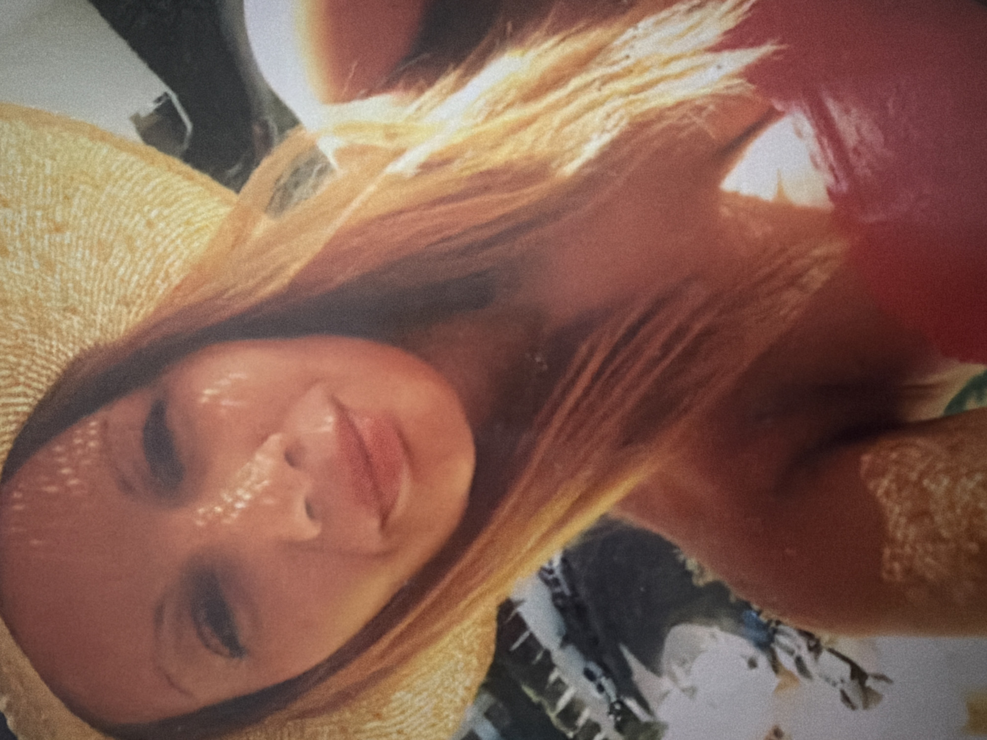 The image depicts a blonde woman wearing a straw hat, with dappled sunlight on her face. She is turned at an angle, with a red object partially visible in the upper right corner. The background is indistinct but appears to include foliage and possibly a building. The location in London, UK is not explicitly discernible from the image.