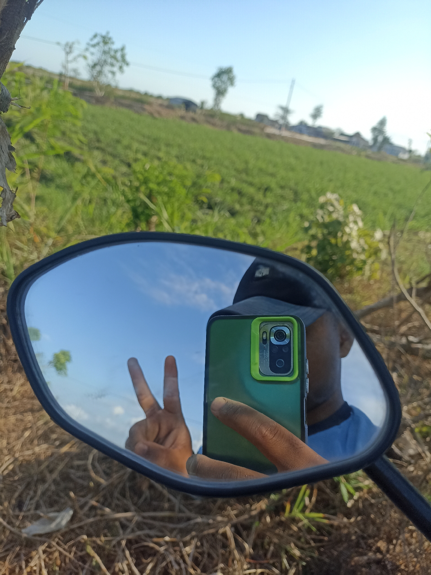 In Pandanarum, Indonesia, a person is taking a selfie in a motorcycle's side-view mirror. The mirror reflects the person's hand holding a green smartphone, making a peace sign, and a portion of their face and blue shirt. Behind the mirror, a grassy field slopes upwards towards a village with houses and trees under a clear blue sky. The foreground consists of dry grass and twigs.