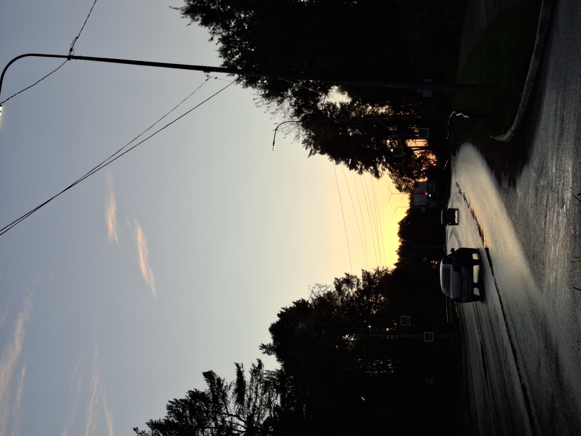 The image depicts a road in the city of Chaikovsky, Russia, at dusk. The sky is a gradient of soft blue and pale orange, with wispy clouds tinged with pink. Silhouetted trees line the sides of the road, creating a dark border against the lighter sky. A streetlamp with its light on is visible on the right, casting an illumination. Several cars are driving on the wet asphalt road, reflecting the fading light. One car, a dark sedan, is prominently featured in the foreground, its rear lights illuminated. In the background, traffic lights show red and green signals. The overall scene suggests a tranquil evening commute on a slightly damp road.