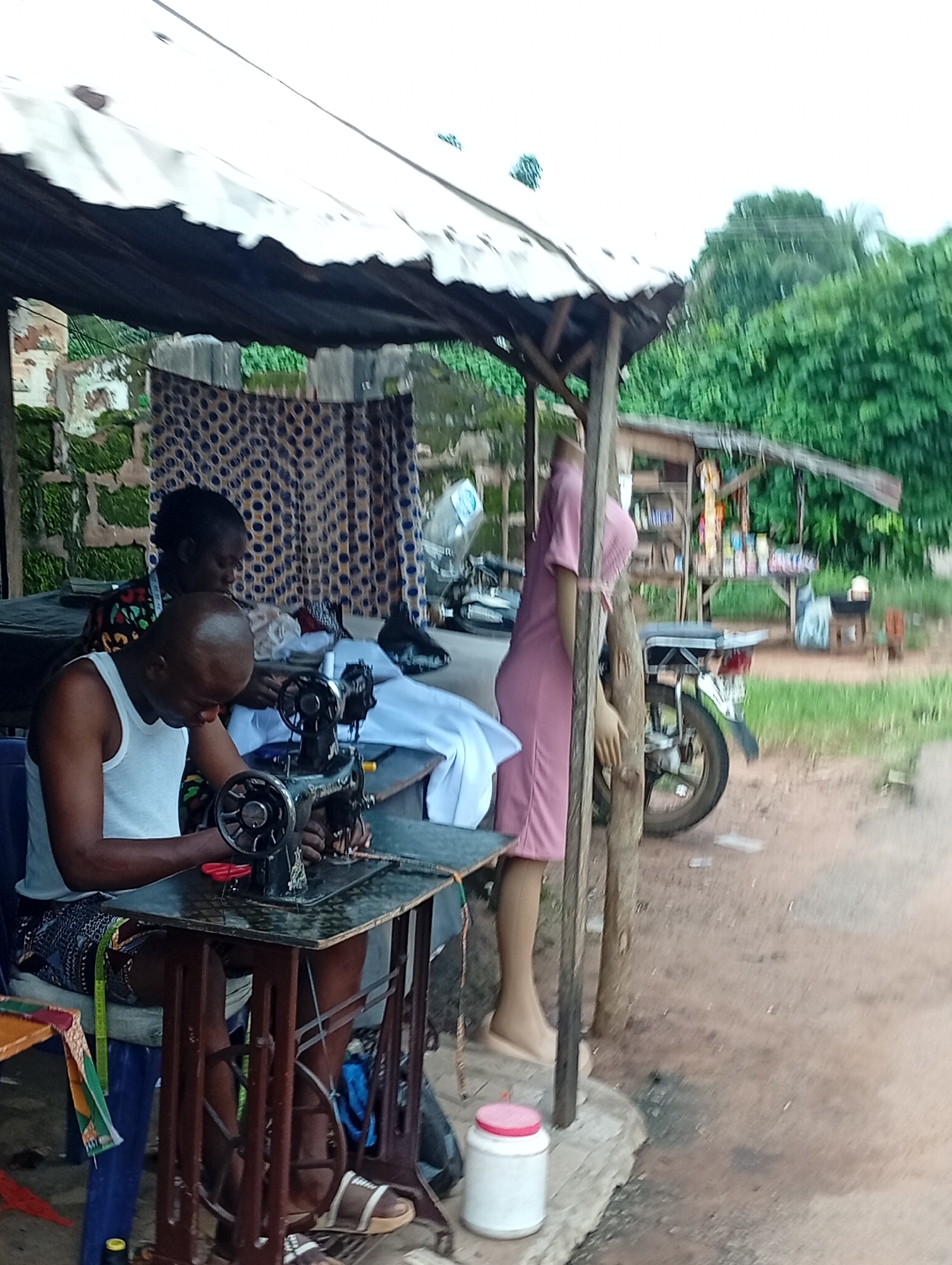 The image depicts a tailoring workshop located in Nsugpe, Nigeria. Two individuals are actively engaged in sewing. In the foreground, a bald man wearing a white tank top is seated at a vintage sewing machine, his hands manipulating fabric. Behind him, a woman with her hair tied back, wearing a patterned top, is also seated, likely assisting or working on a separate task. The workshop appears to be an open-air structure with a corrugated metal roof supported by wooden posts. To the right, a mannequin dressed in a pink outfit stands near a motorcycle. The ground is covered in reddish-brown earth, and the background shows lush green trees and foliage, characteristic of a tropical environment. A small table with various items, possibly goods for sale, is visible further in the background.