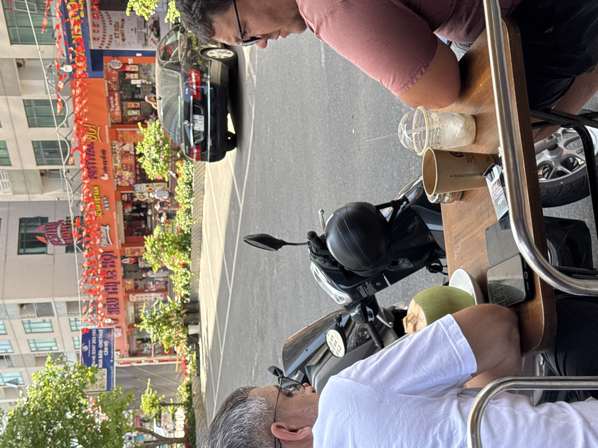 Two middle-aged men sit at an outdoor table in a café. The man on the left wears a white t-shirt and glasses. The man on the right wears a pinkish-red shirt and glasses. The table holds two empty drink cups, a green coconut, a pack of cigarettes, and a cell phone. A motorcycle is parked nearby. Across the street is a multi-story building with hanging red decorations, possibly flags or lanterns. A black car is parked along the street in front of a storefront. The location is Bac Nha Trang Ward, Vietnam.