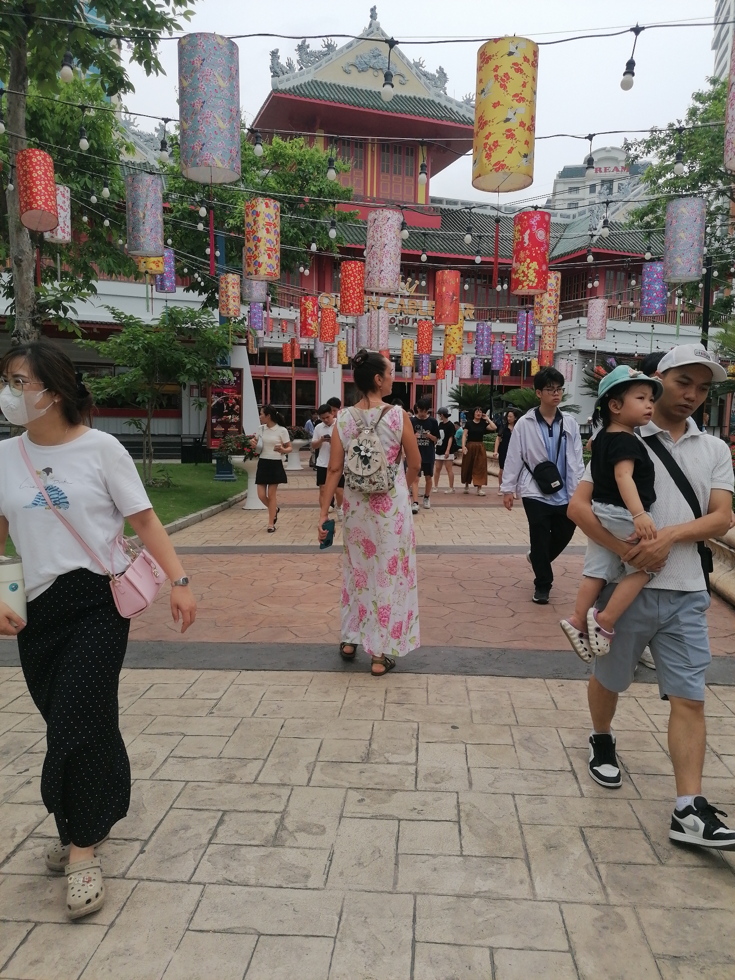 The image shows a street scene in Bai Chay Ward, Vietnam. Many cylindrical lanterns are strung overhead. A group of people are walking along a paved path. A woman in a mask and a black polka dot skirt walks on the left. A woman in a floral dress with a backpack walks in the center. A man carries a child wearing a blue hat. A red and green building with ornate roofs is in the background.