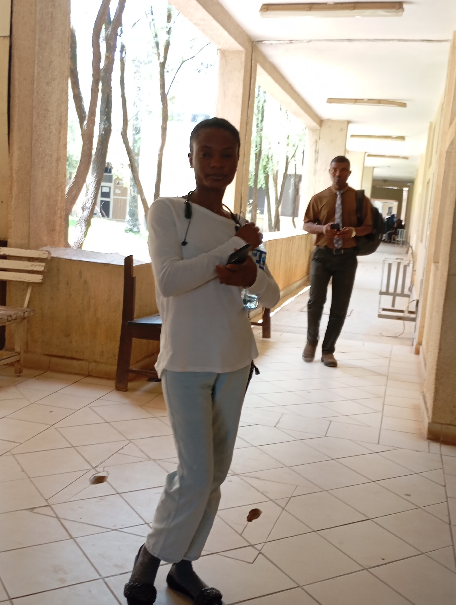 The image depicts a hallway or covered walkway in Gwong, Nigeria. The space is tiled and has large openings to an outdoor area with trees. A woman in a white long-sleeved shirt and light-colored pants stands in the foreground, facing the camera. She has a stethoscope around her neck and is holding a phone or similar device. In the background, a man in a brown shirt and tie walks away from the camera, holding a phone and carrying a backpack. Other individuals are visible further down the hallway, seated. Some chairs are also visible.