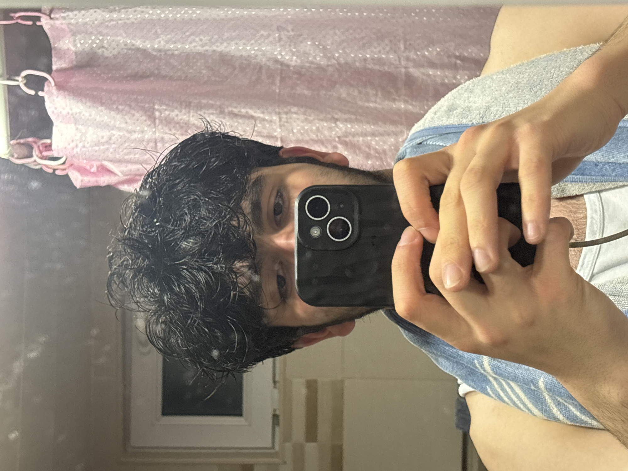 A person is holding a black smartphone with two camera lenses in front of their face, likely taking a selfie in a bathroom mirror. The individual has dark hair and is partially obscured by the phone. A light blue striped towel is draped around their shoulder. In the background, a pink shower curtain and a window with white frames are visible. The location cannot be determined to be Yerevan, Armenia based on the content of this image.