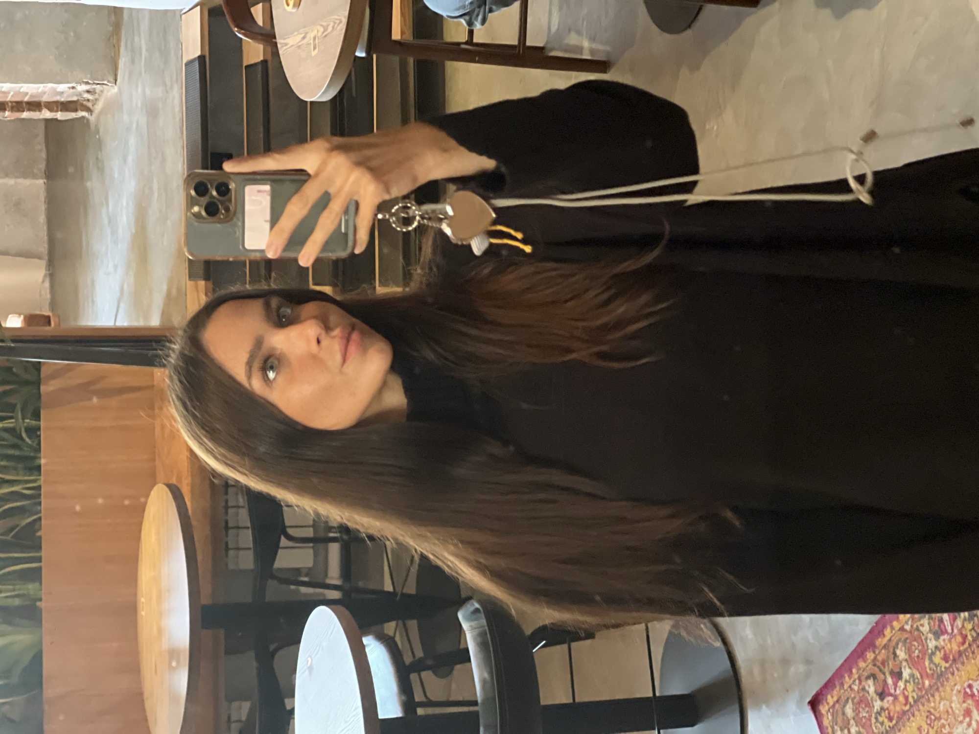 A woman takes a selfie indoors, holding a smartphone. She wears a black top. Her long brown hair has a blonde streak. Tables and chairs are visible in the background, suggesting a cafe or restaurant setting. A keychain hangs from the phone. A patterned rug is visible in the corner. Without additional information, specifying the exact location within Vladivostok, Russia is impossible.