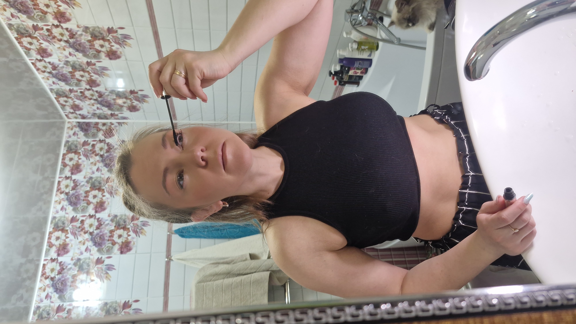 A woman is captured in a mirror selfie, applying mascara to her eyelashes in a bathroom. She appears to be in her 20s or 30s, with light hair, and is wearing a black ribbed crop top and black-and-white patterned shorts or trousers. Her right hand holds the mascara wand to her left eye, while her left hand holds the mascara tube, revealing long, light blue fingernails and a ring on her left ring finger.
The bathroom setting is visible in the reflection, featuring distinctive wall tiles with a floral pattern of pink, purple, and green flowers on a light background. A white sink, a silver faucet, and various toiletries are arranged on a counter behind her. A stack of folded towels hangs nearby. In the upper right corner of the reflection, the head of a fluffy cat is partially visible, observing from the counter. The woman's expression is focused as she applies her makeup. The lighting is bright, suggesting daytime or well-lit artificial illumination. No visible text is present in the image. The scene is set in a bathroom, reportedly in Otchor, Russia.