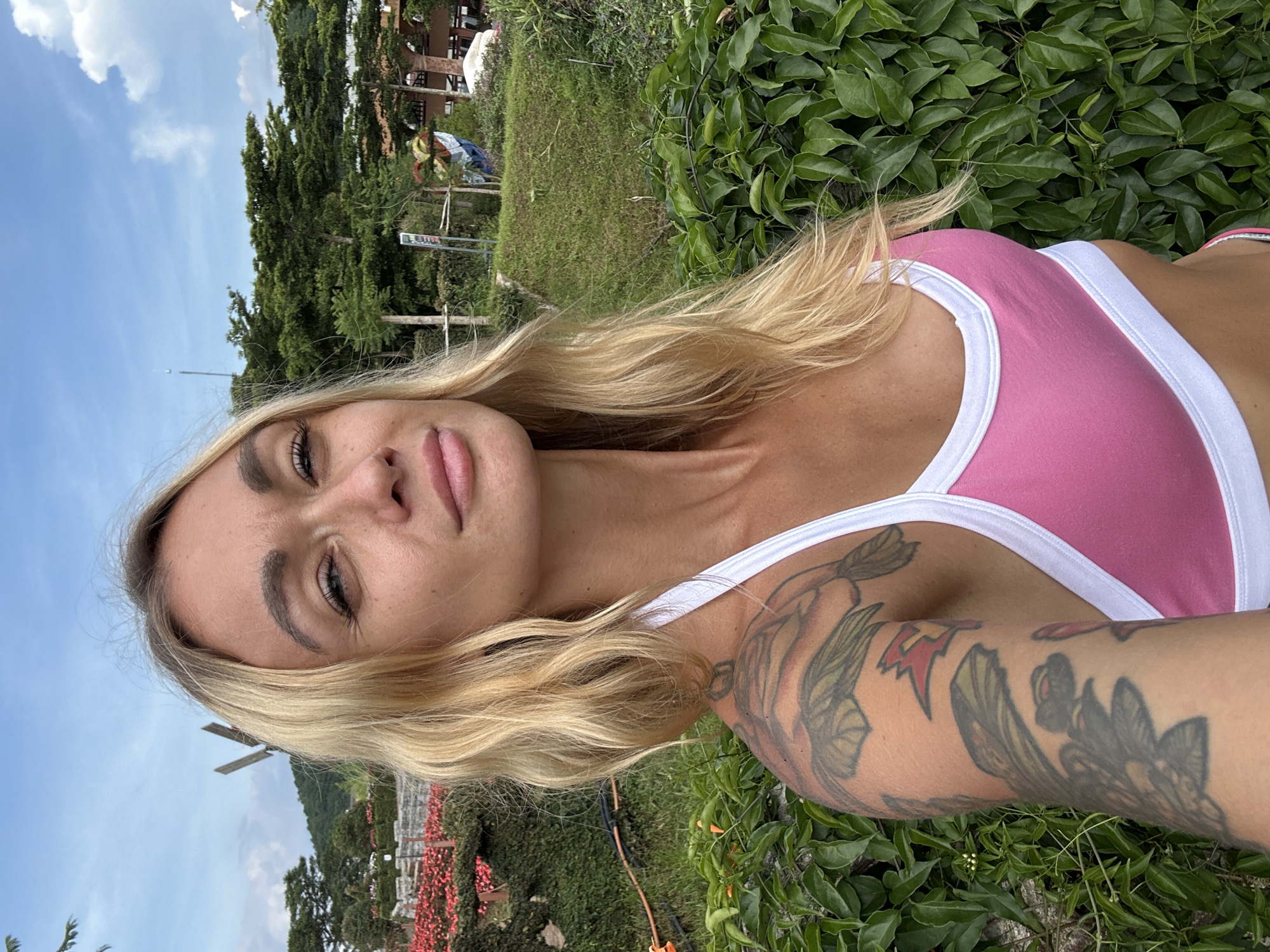 A blonde woman with tattoos on her left arm, wearing a pink and white sports bra, takes a selfie outdoors. She is positioned in the foreground, with lush green foliage behind her and a backdrop of trees, a grassy hill, and a cloudy blue sky. In the distance, there appears to be a garden with red flowers and a small windmill-like structure. The location is Cam Ly - Đà Lạt, Vietnam.