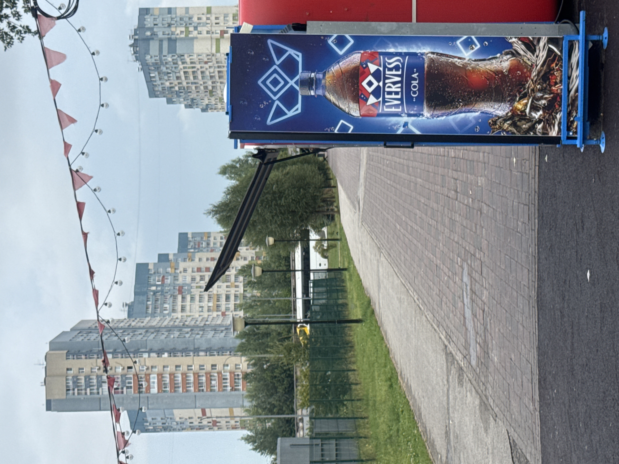 The image shows a view of apartment buildings and a walkway in Nizhny Novgorod, Russia. In the foreground, there is a large advertisement for Evervess Cola displayed on a blue stand. The advertisement features a bottle and glass of cola, with a crown logo. The walkway is paved with bricks and bordered by grass and a fence. In the background, several tall, multi-story apartment buildings are visible, with a cloudy sky overhead. A white bus is seen in the distance, driving on a road behind the fence. String lights with red flags are strung across the top of the image, suggesting an outdoor event or festive atmosphere.