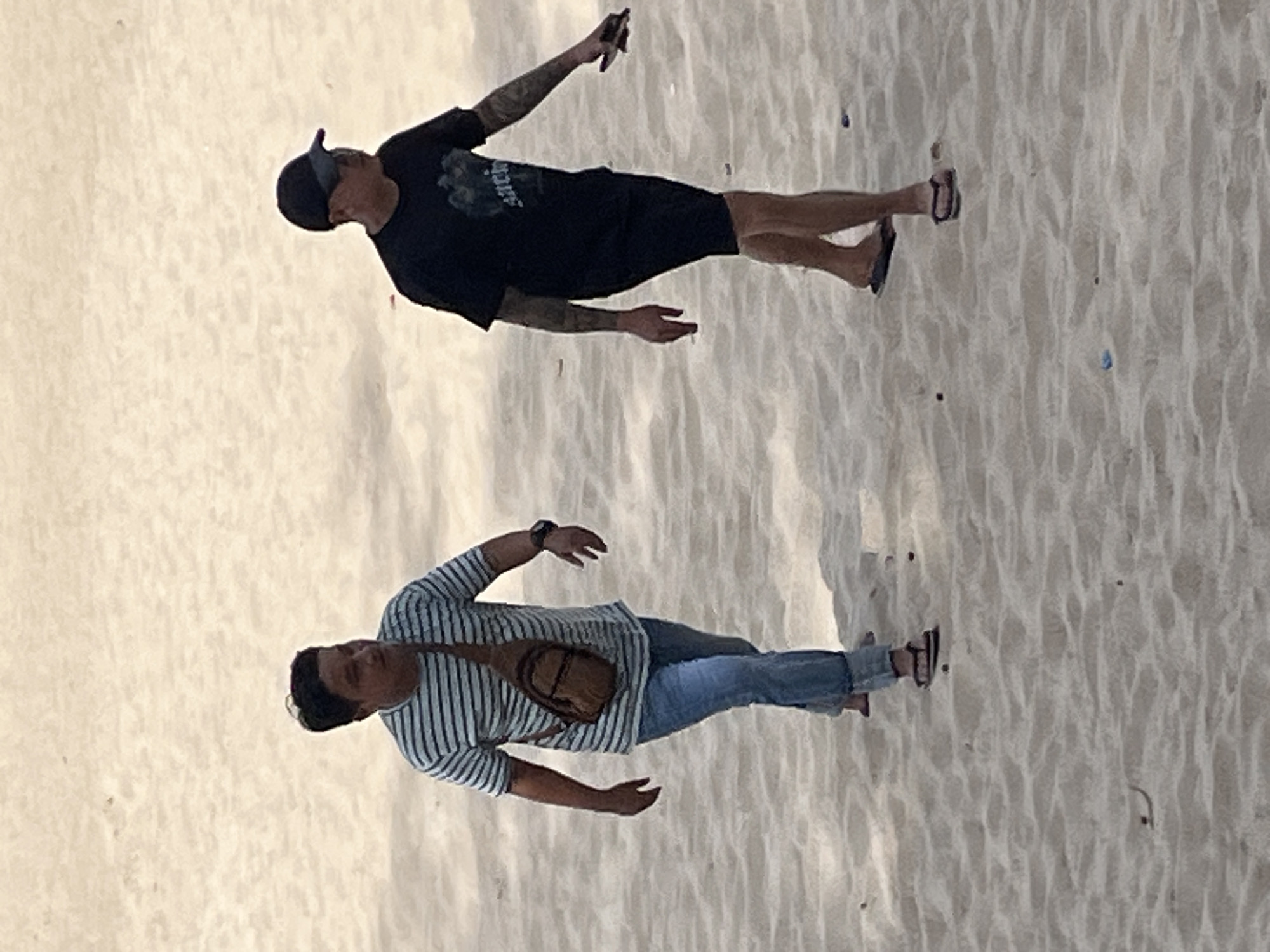 Two men walk on a sandy beach in Mũi Né, Vietnam. The man on the left wears a horizontally striped t-shirt, blue jeans, and sandals, with a brown crossbody bag. He has dark hair and is looking to his left. The man on the right wears a black t-shirt with a graphic design, black shorts, a black baseball cap, and flip-flops. He has tattoos on his arms and is looking to his right, holding a dark object in his right hand. The sand is light brown and appears textured, with some small debris scattered on the surface. The lighting suggests it is daytime.